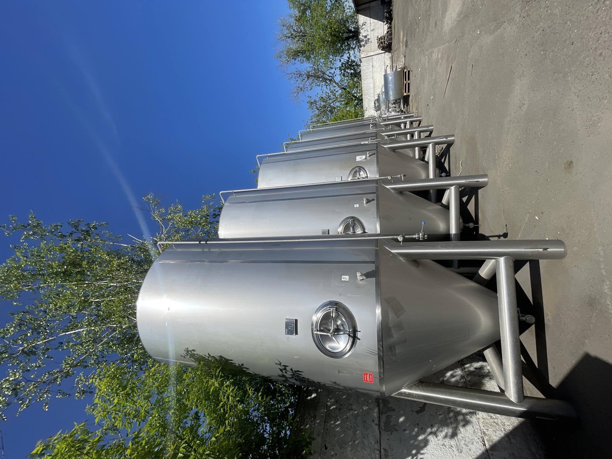 A row of large, gleaming stainless steel conical fermenters stands outdoors beneath a clear blue sky. The tanks are meticulously aligned, receding into the distance, suggesting new equipment awaiting installation or part of an operational facility. Each tank is equipped with a circular manway on its side and is supported by robust metal stands. Bright sunlight casts distinct shadows on the concrete or asphalt ground, indicative of a clear day. Lush green trees are visible around and behind the tanks, providing a natural contrast to the industrial equipment. A concrete building and additional industrial equipment are visible in the background. The foremost tank bears a red rectangular label with white text, possibly "RED" as part of a brand, located below its manway. No human activity is depicted. The scene suggests an outdoor industrial facility, likely a brewery or beverage production plant, in Krasnoyarsk, Russia.