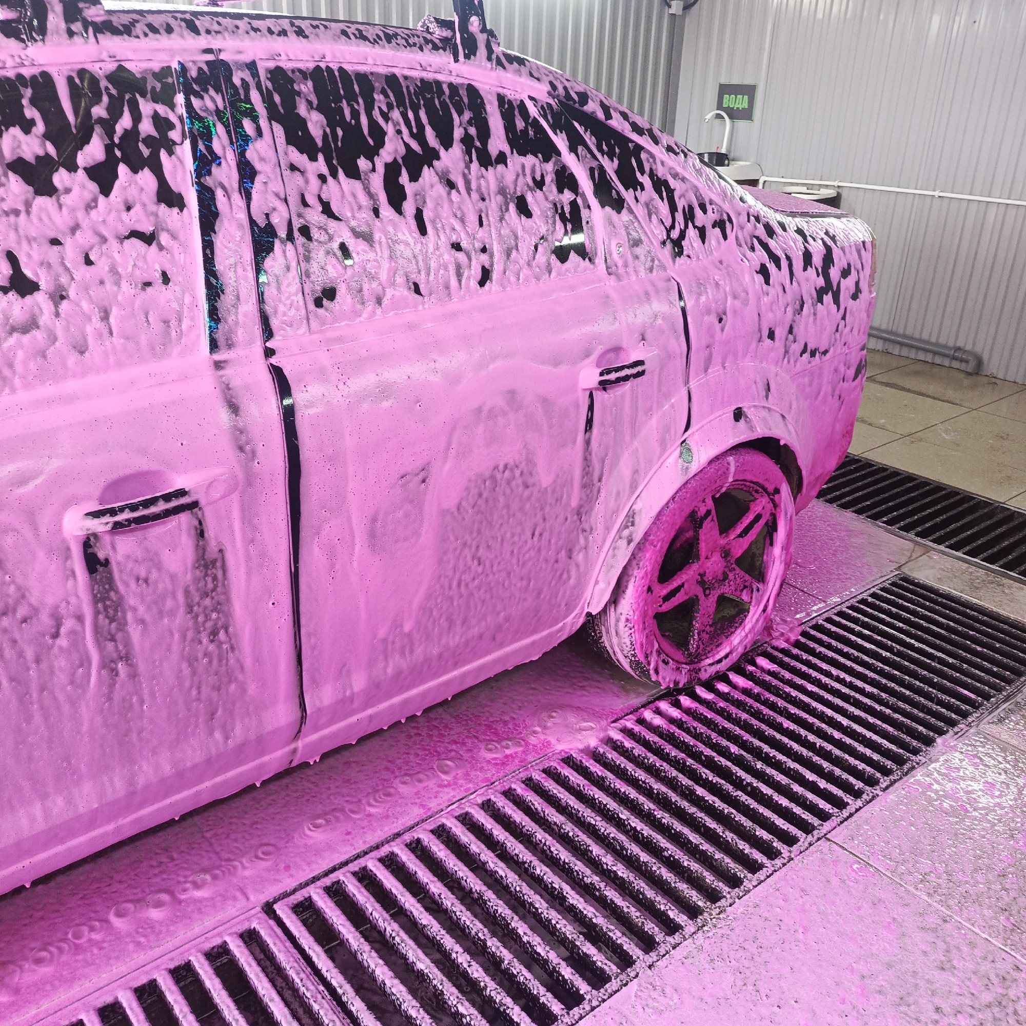 This media file captures a dark-colored car, likely a sedan, covered in vibrant pink washing foam inside what appears to be a car wash bay in Chaikovsky, Russia. The car is seen from the side, angled slightly towards the rear, with the driver's side facing the viewer. Thick pink foam blankets most of the vehicle's body, including the rear wheel and parts of the roof rack. The foam cascades down the sides, collecting on the floor and into large metal drainage grates running across the foreground.
Through gaps in the foam, the dark color of the car is visible, particularly on the window frames and handles. Some windows show dark reflections, while one crop reveals iridescent, multi-colored reflections, possibly from a holographic sticker or unique window tint. The rear wheel, completely enveloped in foam, has a distinct multi-spoke rim design.
The setting is an indoor space with corrugated metal walls. In the background, to the left of the car's rear, a white faucet and basin are visible. Above them, a green sign with white lettering clearly displays the Russian word "ВОДА," meaning "WATER." The tiled floor of the car wash bay is partially covered in pink foam and water, flowing into the prominent drainage grates. The scene depicts an active stage of a car wash, highlighting the aesthetic of the brightly colored cleaning foam.