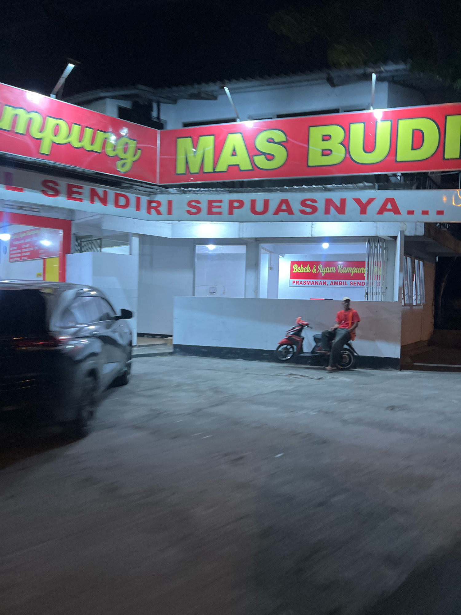 The image depicts a nighttime scene outside a restaurant in Tempel, Indonesia. A large, bright red sign with yellow text, partially obscured, reads "MAS BUDI" in large letters. Below it, another sign in red text says "SENDIRI SEPUASNYA...". The restaurant appears to be an open-air establishment with white walls. Inside, a smaller red sign reads "Bebek & Ayam Kampung" with further text below indicating "PRASMANAN, AMBIL SENDIRI". Two people are visible: one is sitting on a red motorcycle to the right of the frame, wearing a light-colored cap and an orange shirt, and dark pants. Another person is seated on the same motorcycle. To the left, a portion of a dark-colored SUV is visible, with its headlights on. The ground is paved, and the overall lighting suggests it is nighttime.