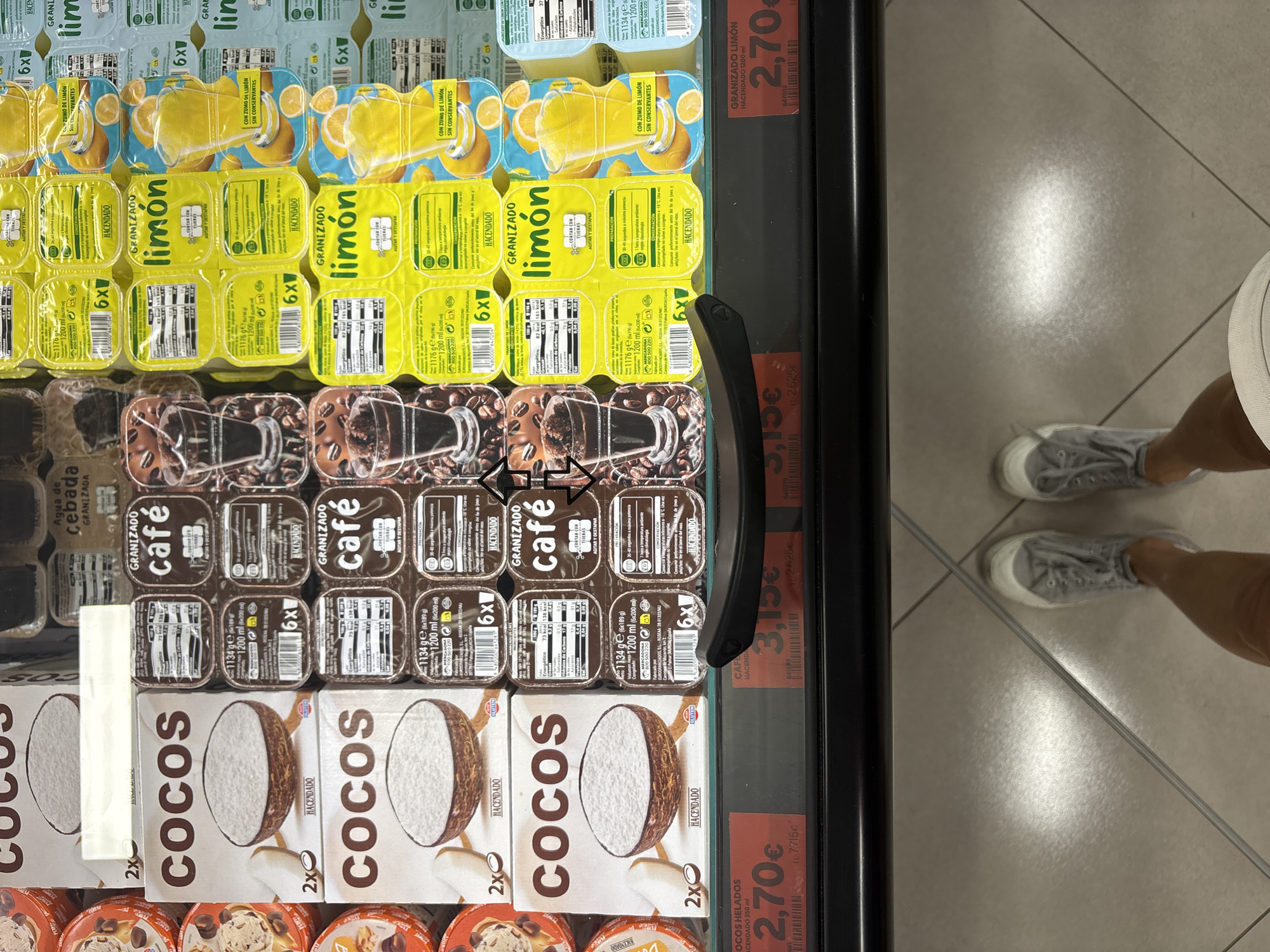 The image shows a person's legs and feet wearing grey canvas sneakers, standing on a tiled floor. Above the floor, a close-up view of a supermarket freezer display is captured. The display is stocked with various frozen desserts. Prominently featured are packages of "COCOS" ice cream, described as "GRANIZADO" with coconut, sold in packs of two. There are also packages of "GRANIZADO café" (coffee flavor) and "GRANIZADO limón" (lemon flavor). Each of these granizado products is sold in a pack of six individual cups. Price tags are visible below the freezer display, indicating prices of "2,70€" for "Cocos Helados" (frozen coconut), "3,15€" for "Café" (coffee), and "2,70€" for "Granizado Limón" (frozen lemon). The overall scene suggests a shopping trip in a supermarket in València, Spain, likely a Mercadona supermarket given the product branding "Hacendado."