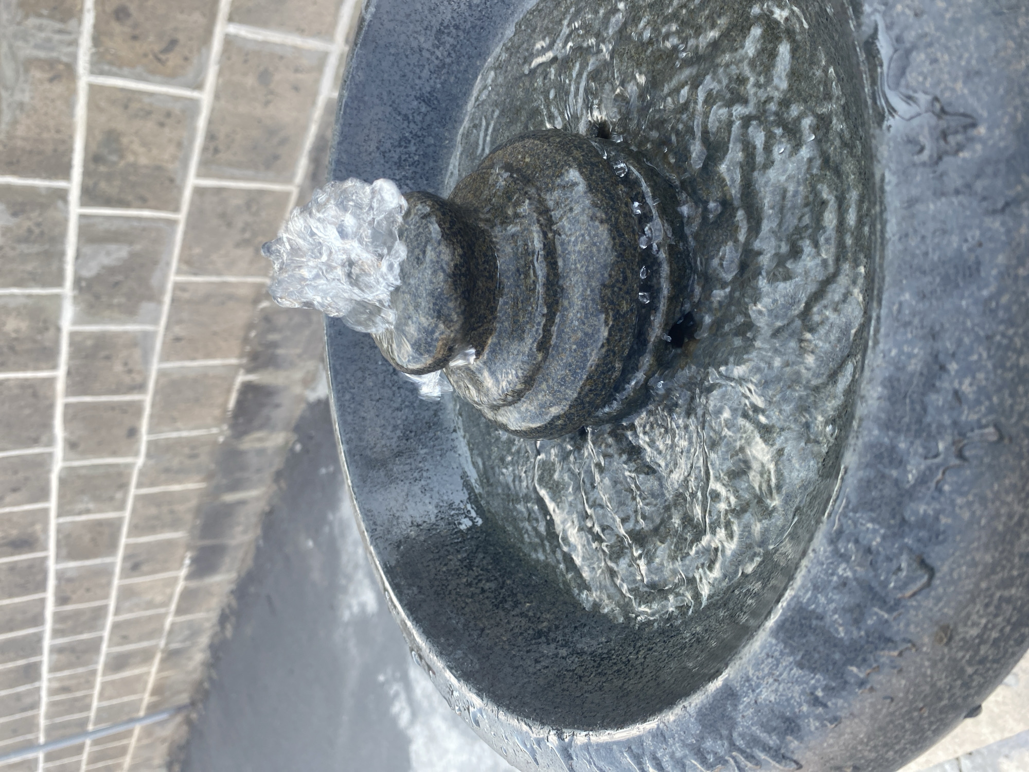 This is a close-up, eye-level, medium shot of a stone fountain in a city environment. The fountain is made of dark gray, rough-textured stone and is filled with water. A small, decorative stone structure sits in the center of the fountain, from which a stream of water sprays upwards, catching the light. The water in the fountain is rippling and shows a texture of small waves. The exterior of the fountain bowl also shows droplets of water. Behind the fountain, a wall constructed of rectangular, light-colored bricks is visible, with a gray paved surface in front of it. The overall lighting suggests an outdoor, daytime setting. There are no people visible in the image.