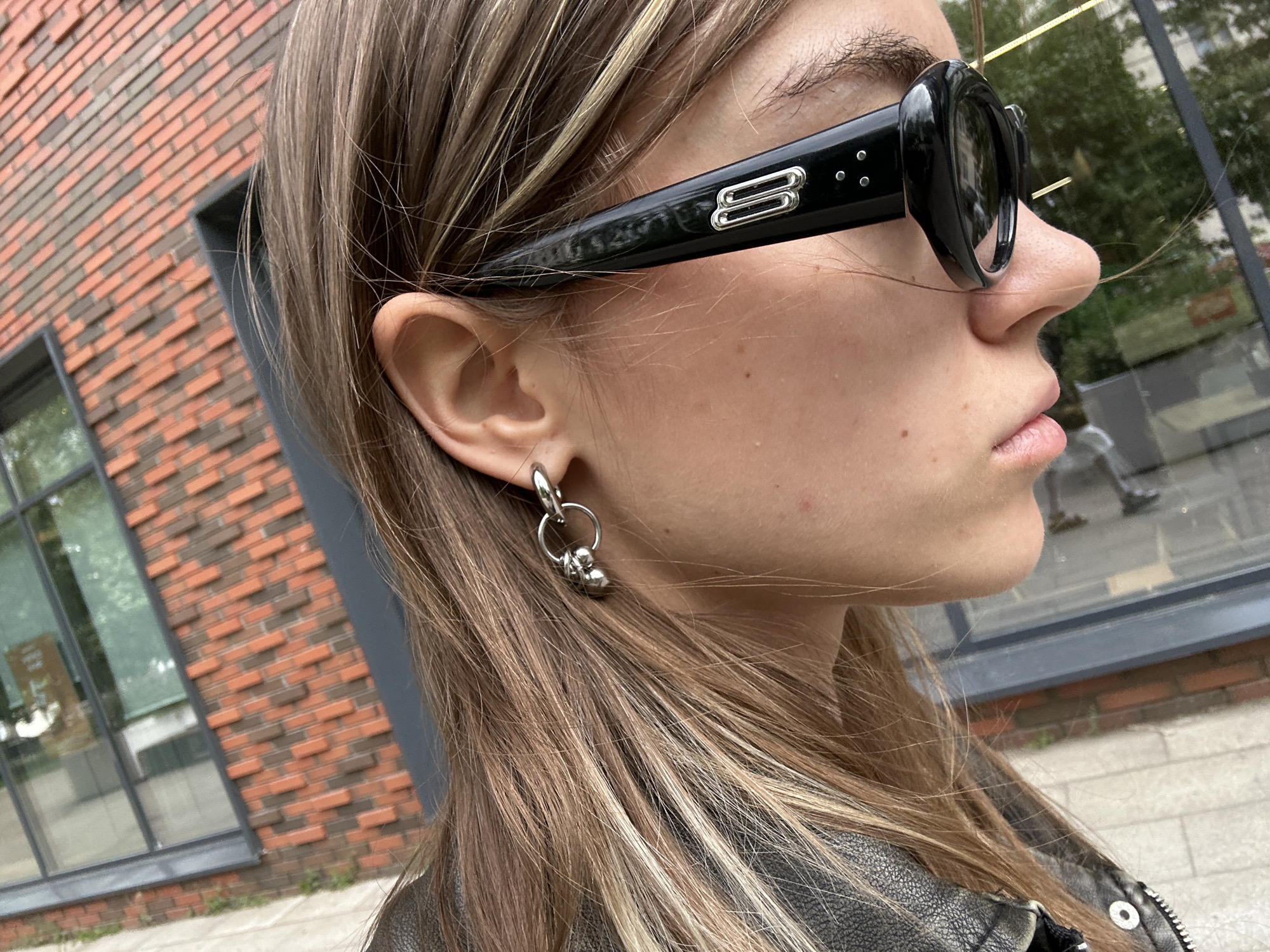 A close-up shot captures the profile of a young person wearing large, black, oval-shaped sunglasses with a silver emblem on the left temple. They have long, blonde-streaked brown hair that falls over their shoulder and are wearing a silver hoop earring with two small spherical charms. The person is wearing a black leather jacket. In the background, a brick building with a modern glass facade is visible, suggesting an urban environment. The lighting indicates it is daytime, and the reflection in the window suggests the presence of other people. This image does not contain information about the specific location of Moscow, Russia.