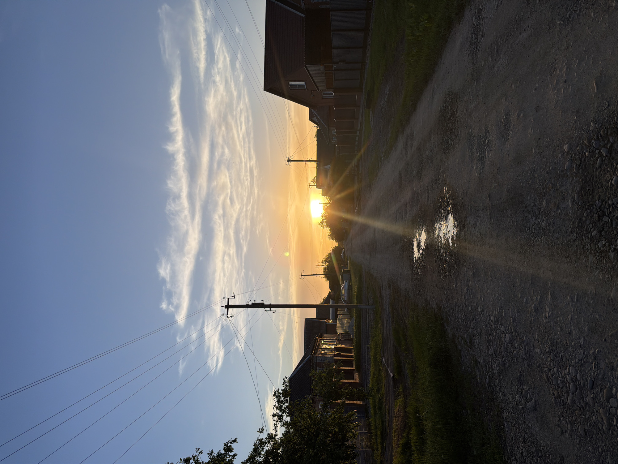 The image depicts a rural road lined with houses at sunset. A dirt road stretches into the distance, reflecting sunlight in a puddle. The sun is positioned low on the horizon, between the houses, casting a golden light. Power lines run parallel to the road, supported by wooden poles. Clouds are visible in the sky. The location is likely in Leningradskaya, Russia, based on external information, characterized by its rural landscapes and residential architecture.
