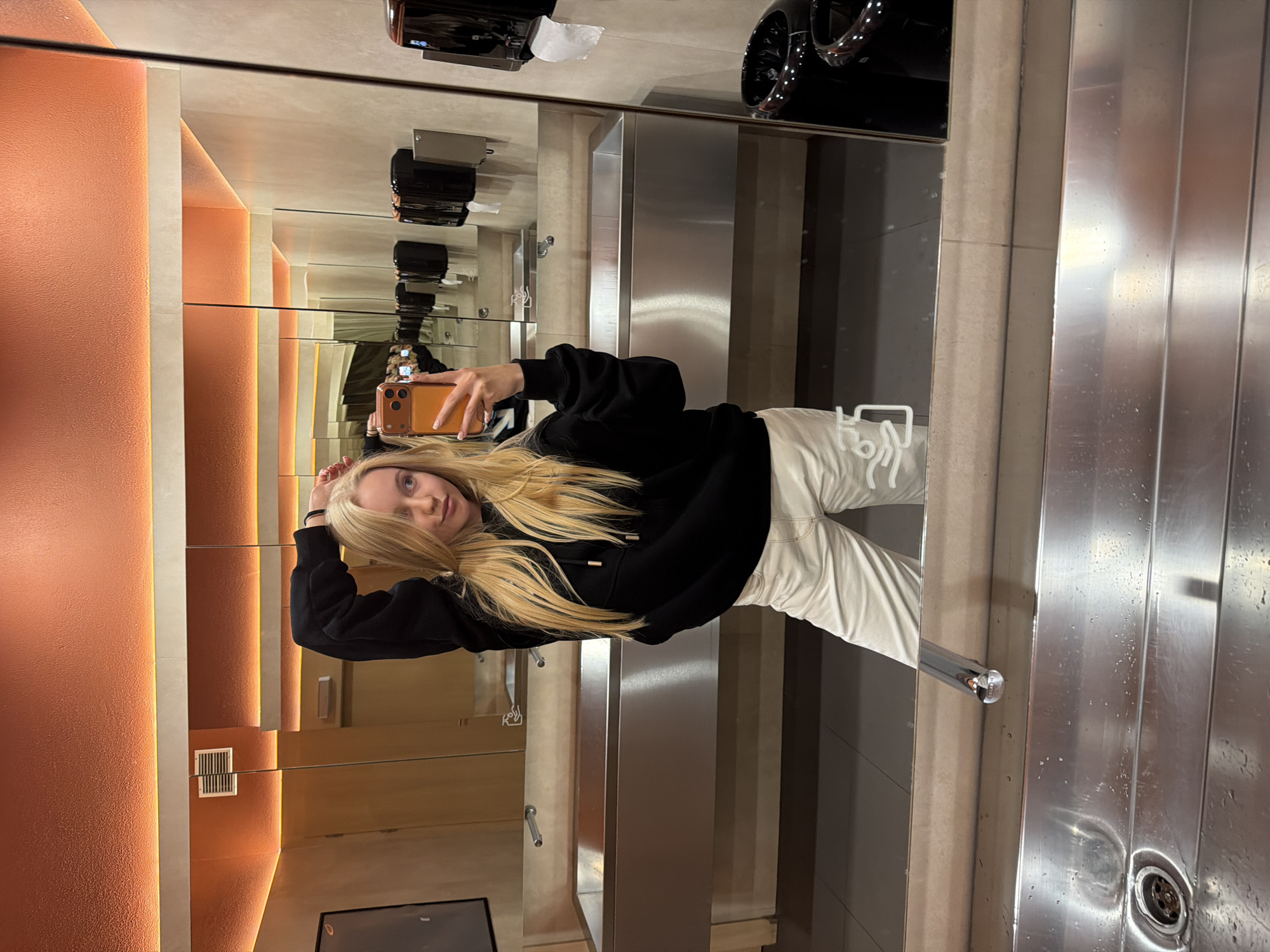 A young woman is taking a selfie in a bathroom. She is wearing a black hoodie and white pants. She is holding her phone in her right hand. The bathroom has a stainless steel sink, an orange wall, and a mirror. There are paper towel dispensers and hair dryers above the mirror. The location is Unknown, Unknown.