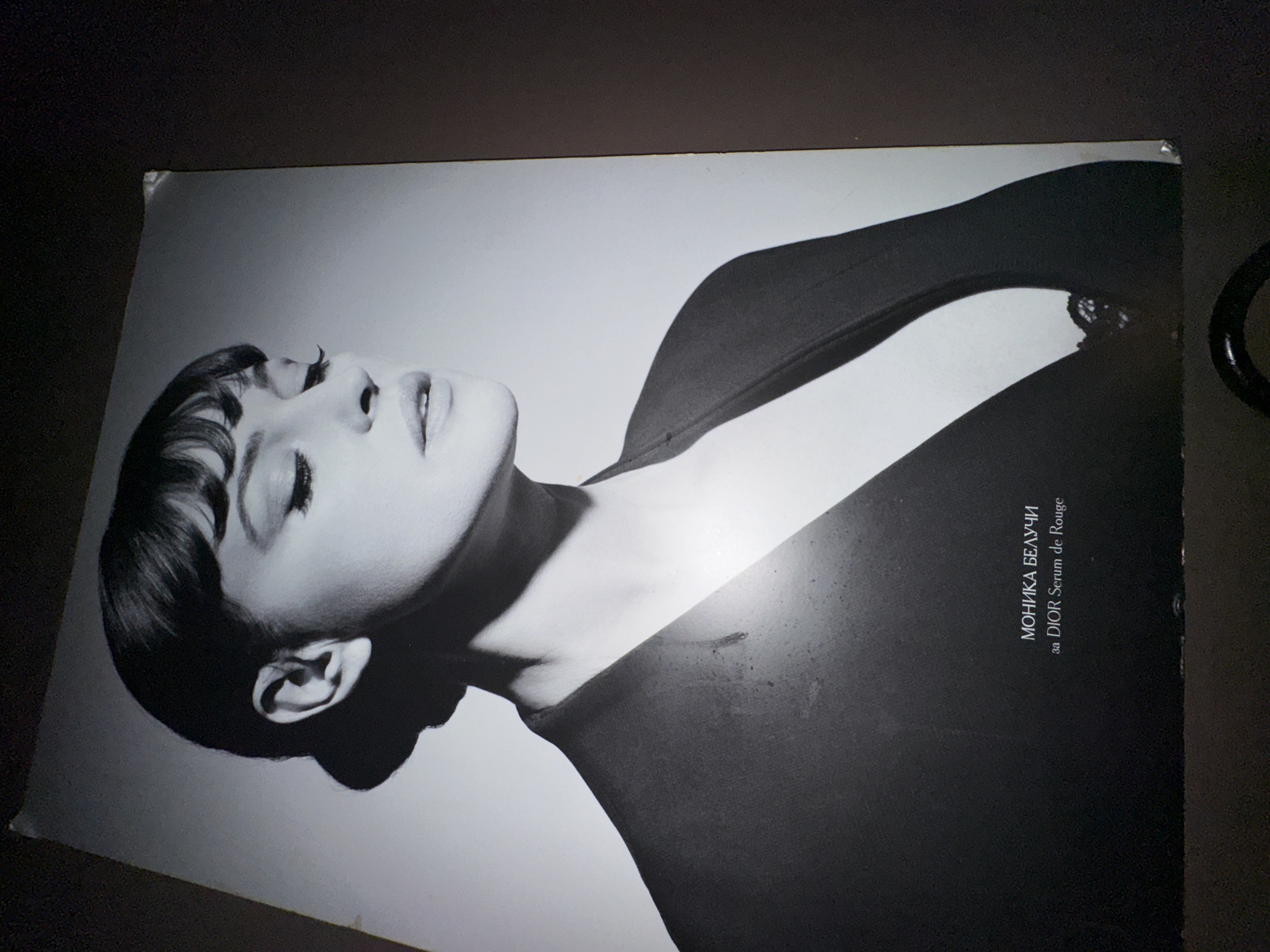 A black and white poster shows a woman with a dark, short haircut looking up. Text in Cyrillic script on the right reads "МОНИКА БЕЛУЧИ за DIOR Serum de Rouge". The poster appears to be lying on a dark surface. There is no evidence to indicate this poster is located in Sofia, Bulgaria.