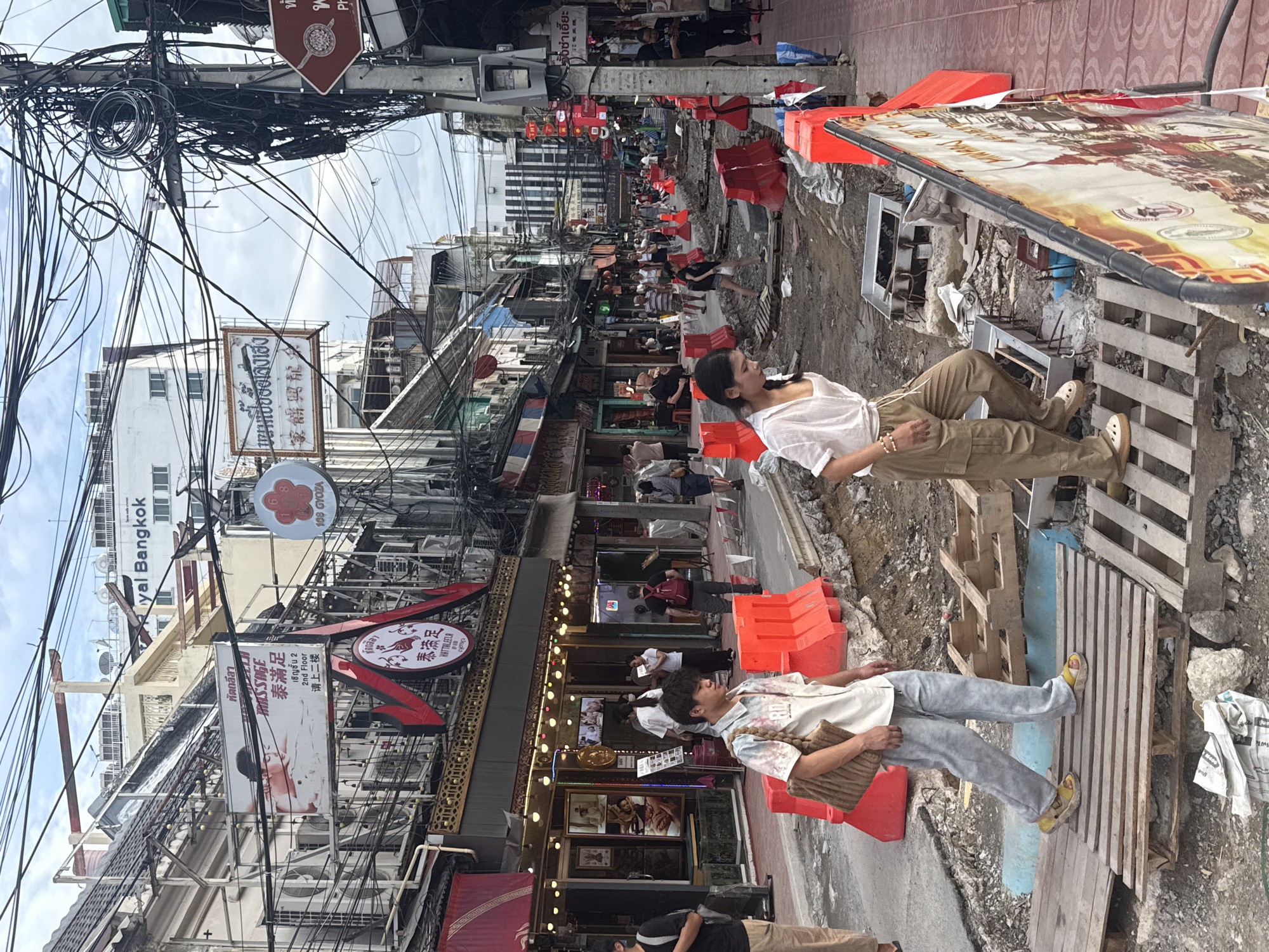 This image captures a street scene in Bangkok, Thailand, likely in the Yaowarat (Chinatown) area, characterized by its dense urban environment and ongoing construction. In the foreground, two individuals are walking on makeshift wooden platforms over a construction site that has disrupted the sidewalk and street. The man on the left, wearing a patterned short-sleeved shirt and light-colored pants with sandals, carries a large brown bag. The woman on the right wears a white loose-fitting shirt and khaki cargo pants.
The background reveals a bustling street lined with multi-story buildings adorned with numerous signs, many in Thai and Chinese, indicating shops and businesses, including massage parlors and restaurants. A prominent sign says "Royal Bangkok." The scene is further complicated by an extensive network of overhead electrical wires and utility poles, a common sight in Bangkok. Orange construction barriers delineate the work area, with piles of dirt, rubble, and construction materials visible. Pedestrians can be seen further down the street, navigating around the construction. The sky is overcast.
