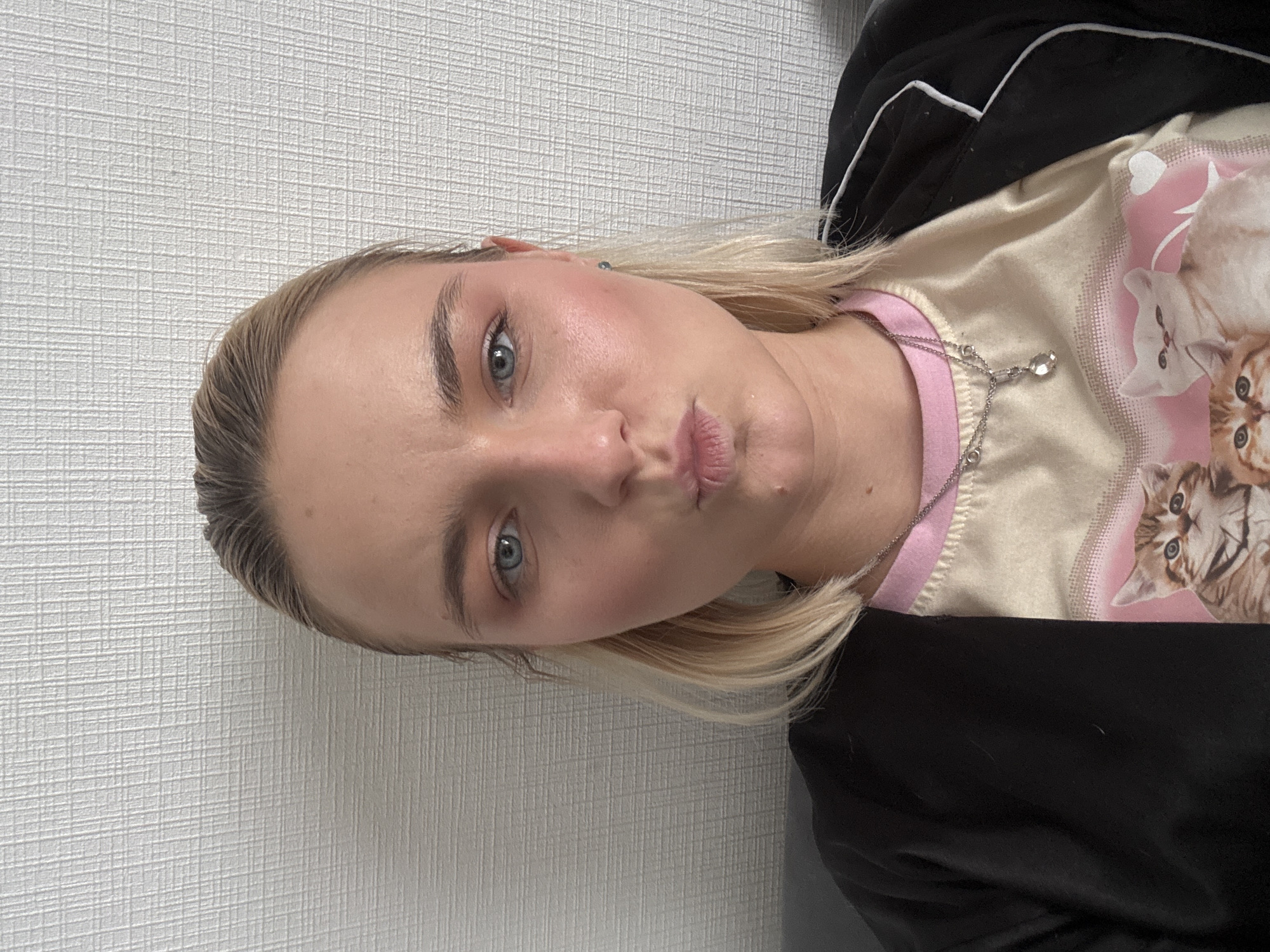 A young woman with light blonde hair and blue eyes is posing for a selfie. She is indoors with a textured white wall as a backdrop. The woman is wearing a black top with white piping and a beige t-shirt with a pink collar and a cat print. She is also wearing a silver necklace. Her lips are pursed as if she is blowing a kiss. The location is Unknown, Russia.