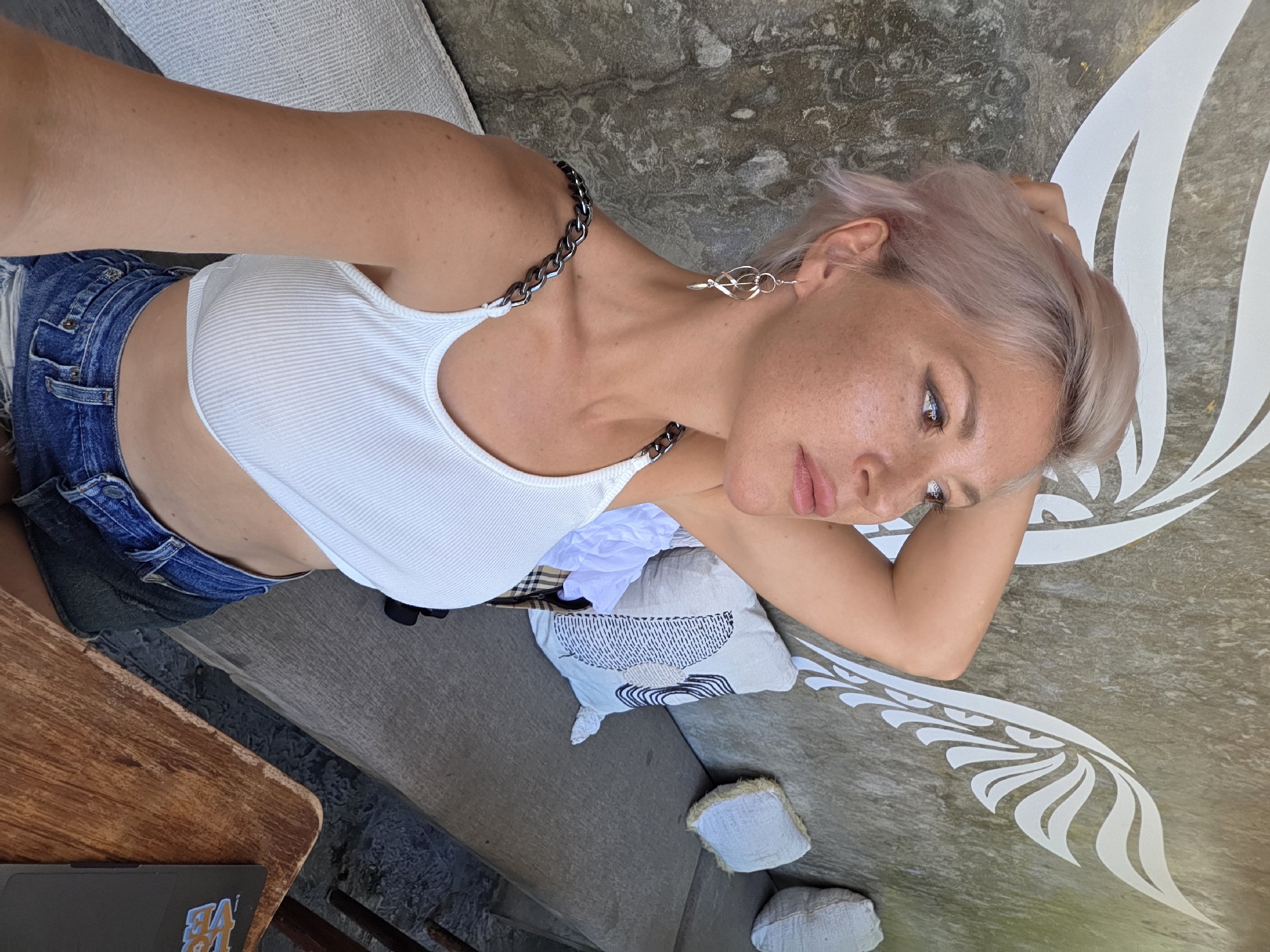 A young woman with short, light pink hair lies on a stone-like surface, possibly a bench or platform, that is decorated with a white painted design resembling stylized wings or leaves. She wears a white ribbed tank top, denim shorts, and silver earrings, with a silver chain strap or necklace. Her gaze is directed slightly downward and to the right of the frame, and her arm is bent as it supports her head. Behind her, a collection of pillows and a white cloth are visible. A wooden table with a laptop sits at the edge of the frame. The scene is situated outdoors in North Kuta, Indonesia.