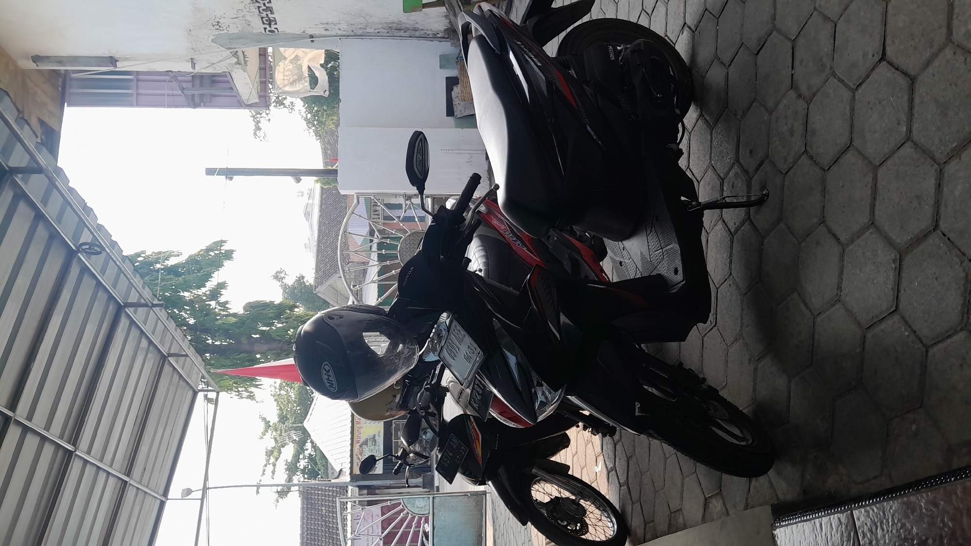 Two parked motorcycles are visible in the foreground, with a black motorcycle in front and a black and red motorcycle behind it. A black helmet with an "NHK" logo is placed on the handlebars of the front motorcycle. The motorcycles are parked on a hexagonal paver-stone surface under a metal awning. In the background, there are buildings and trees, and a red and white flag is visible. The scene appears to be outdoors in a street or parking area in Sambong, Indonesia, possibly near a shop or restaurant given the awning and the presence of what looks like a business sign in the background. The lighting suggests it is daytime.
