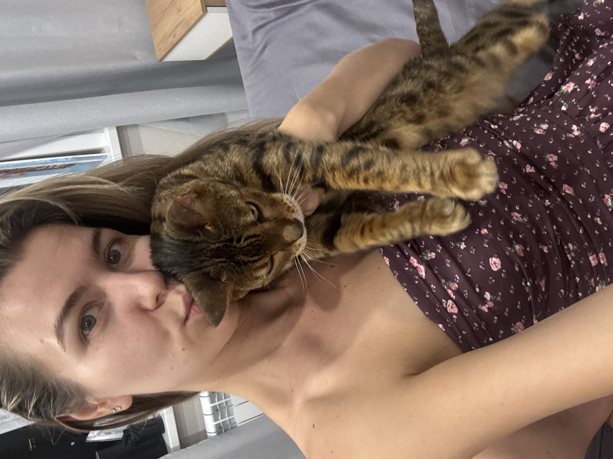 In an indoor setting, a young Caucasian woman with long blonde hair holds a tabby cat. The woman, facing forward with her mouth puckered, is wearing a strapless dark purple dress with a floral print. The cat, nestled against her chest, is looking down with its eyes partially closed. The background includes a grey curtain, a wooden bedside table with a white drawer front, and a white radiator. The lighting suggests an indoor environment, possibly a bedroom or living room.
