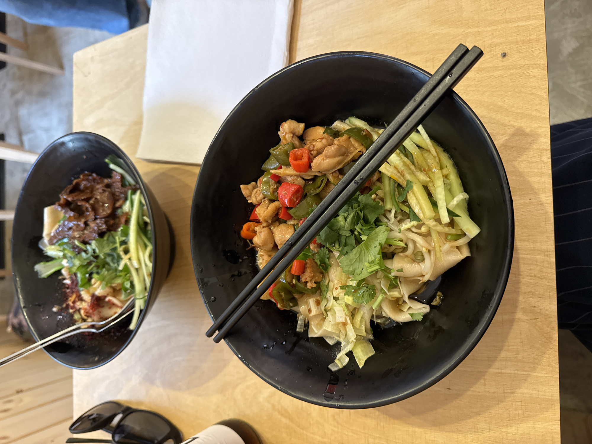 The image shows two black bowls of Asian cuisine sitting on a wooden table. The larger bowl in the foreground contains noodles, chicken, red and green peppers, cilantro, and what appear to be julienned cucumbers. A pair of black chopsticks rests diagonally across the top of this bowl. The smaller bowl to the left contains meat (possibly beef), cilantro, and more of the julienned cucumber. A fork is visible in the smaller bowl. Below the smaller bowl, partially cropped out, are sunglasses and a white bottle. The table has a light wood grain. The background shows glimpses of other furniture, including a chair and a white paper napkin. No information about the location in Sofia, Bulgaria is available from the image.