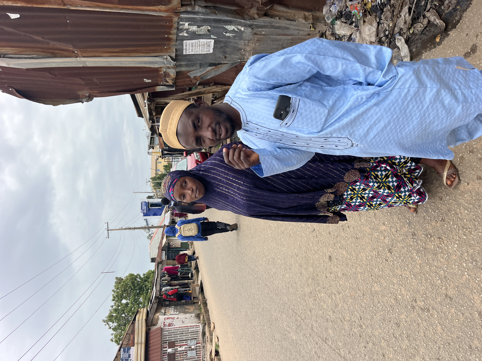 A medium shot captures a Muslim man and woman standing on an unpaved street in what appears to be a market or residential area. The man, on the right, wears a light blue traditional outfit with a tan kufi cap. A smartphone is visible in his chest pocket. He gestures with his right hand towards the camera. The woman, to his left, wears a dark blue hijab and a patterned dress. They are positioned in front of a corrugated metal structure, with a street stretching into the background lined with shops and a few other pedestrians. The sky is overcast.
