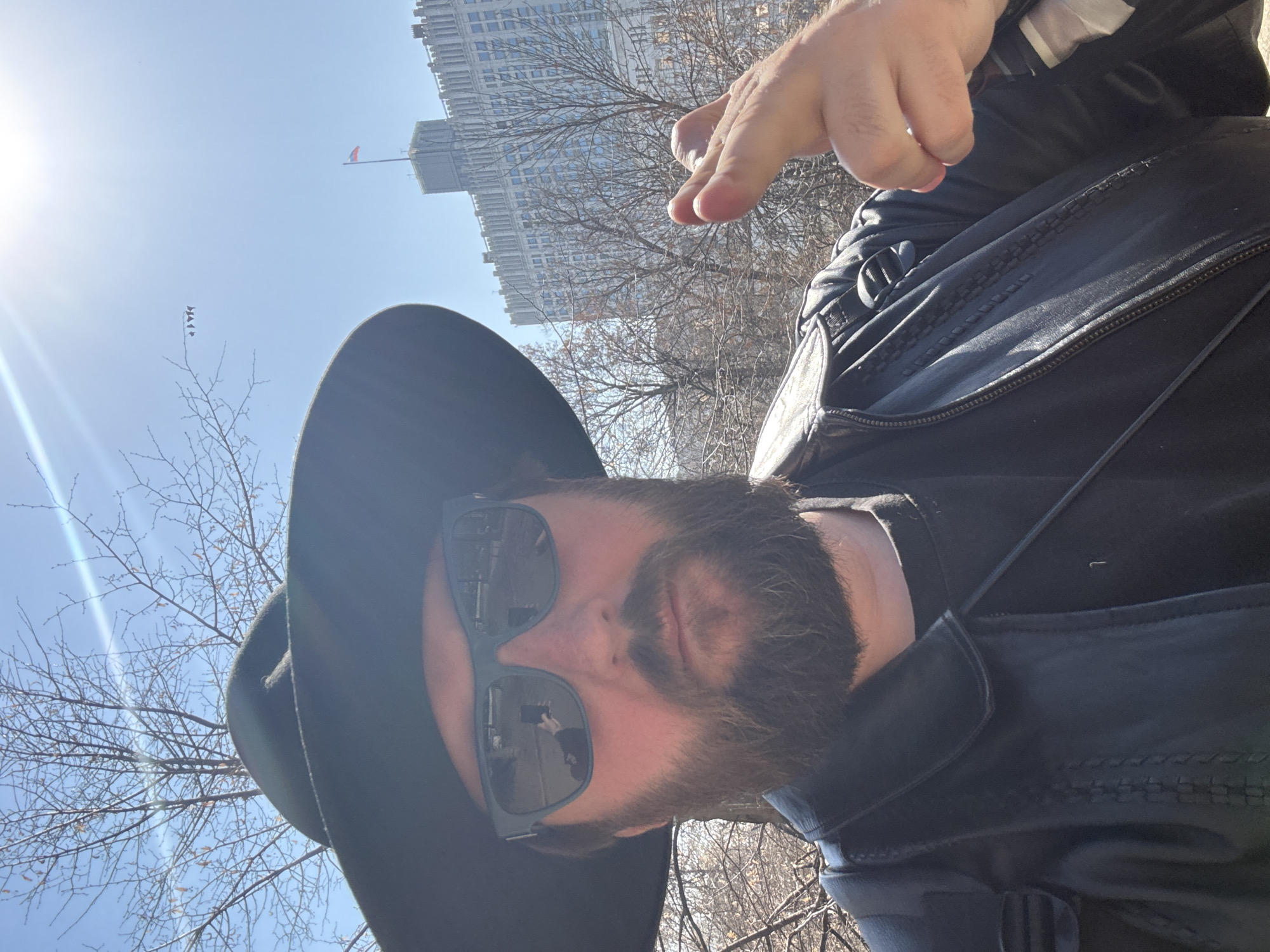 The image depicts a man in a black hat, sunglasses, and a leather jacket, making a hand gesture in an outdoor setting. Behind him, through bare trees, is a large, imposing building that is identified as the Hotel Ukraina, one of Moscow's "Seven Sisters" Stalinist skyscrapers. The scene is set during daylight hours, with a clear sky and sunlight present.