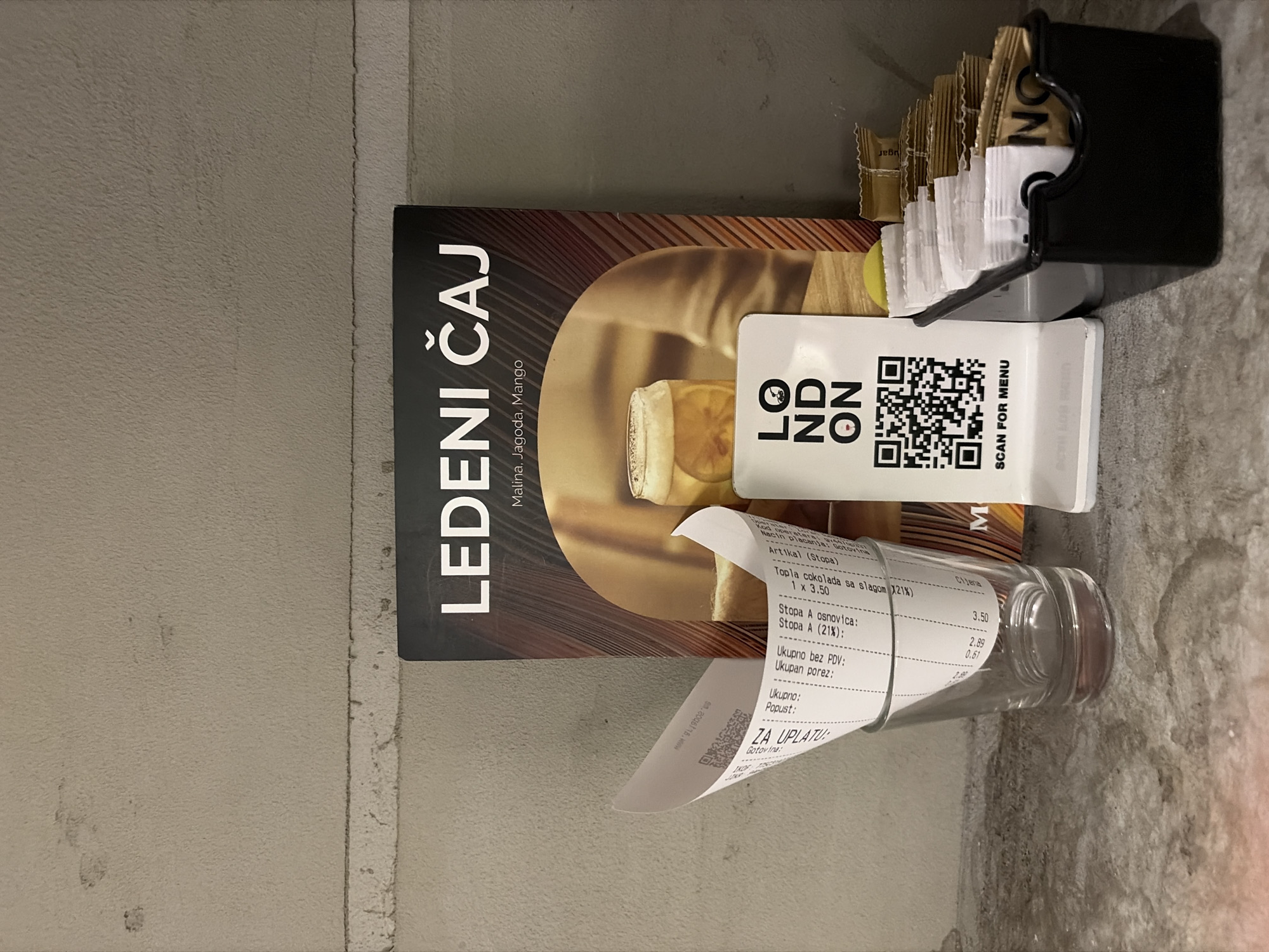 The image shows a collection of items on a surface, likely a table or countertop. There's a menu or advertisement for "LEDENI ČAJ" (Iced Tea) with flavors Malina (raspberry), Jagoda (strawberry), and Mango. On top of the menu, there's a small card with the "LON ODN" logo and a QR code labeled "SCAN FOR MENU". Beside the card, there is a black container holding sugar packets. Resting on the surface is a clear glass with a partially visible receipt wrapped around it. The receipt itemizes an order including "Topla cokolada sa slagom (21%)" (Hot chocolate with cream 21%).
Given the name of the drink and location information in the OCR, it is possible that this image may have been taken in Budva, Montenegro.