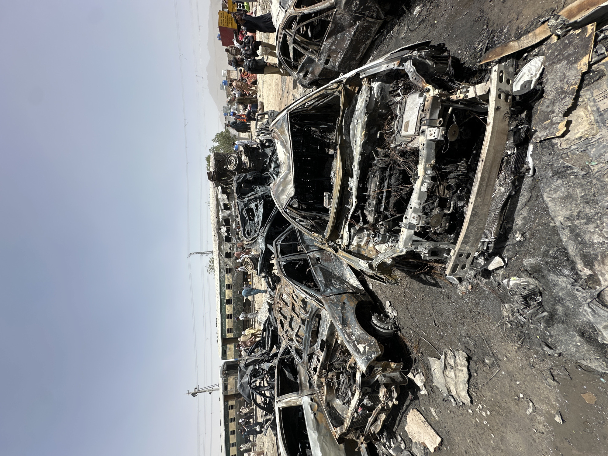 The image shows a scene in Quetta City Tehsil, Pakistan, depicting a graveyard of burned-out cars. The vehicles are reduced to charred frames, with exposed wires and components. In the background, a train and buildings are visible, along with a group of people observing the scene. The environment appears dry and dusty.