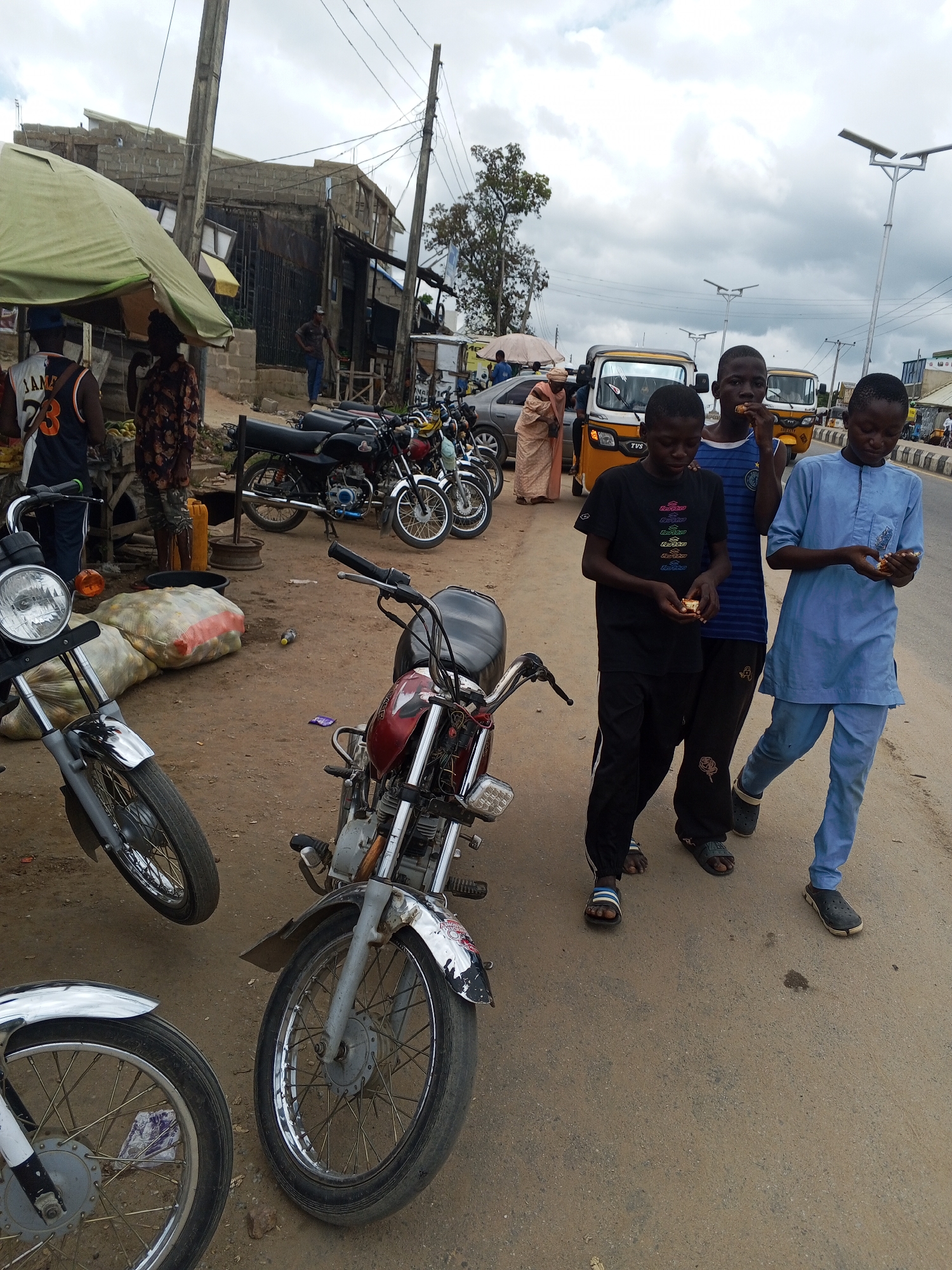 In Zango-ndaji, Nigeria, on a cloudy day, a street scene unfolds with several motorcycles parked on a dirt and asphalt road. Three young males are the central figures, walking and eating. One boy, wearing a blue and white striped t-shirt and dark pants, is in the middle, eating a piece of food. To his left, a boy in a black t-shirt and black pants also holds and eats a piece of food. To the right, another boy in a light blue traditional outfit and matching pants is walking and holding food.
Behind them, several motorcycles are parked. Two are prominent in the foreground, one silver and black with a chrome fender, and a red and silver motorcycle with visible engine parts. In the background, more motorcycles are lined up. A yellow and white tricycle is also visible.
The environment includes a roadside stall with a large green awning, partially obscuring a building constructed from cinder blocks. Bags of produce, possibly yams, are stacked near the stall. Electrical poles and wires crisscross the sky, indicating urban infrastructure. The road is dusty, with patches of asphalt. The general atmosphere suggests a bustling local market or transportation hub.