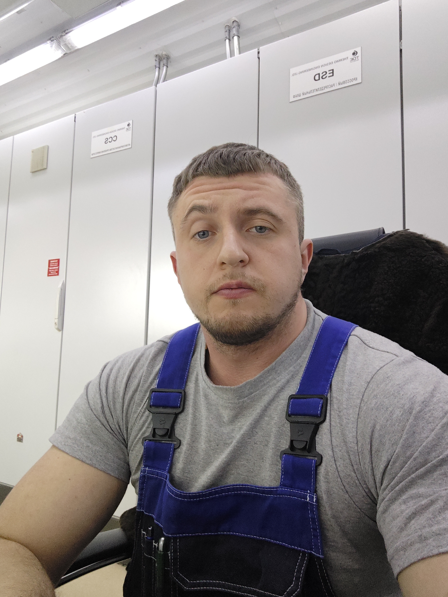 A man with short, light brown hair and blue eyes is centered in the frame, looking directly at the camera with a neutral expression. He has a beard and appears to be wearing a gray t-shirt with dark blue overalls. The man is sitting in an office chair, and part of the dark, furry backrest is visible behind his right shoulder.
The background consists of white, modular-looking cabinets or lockers. On the upper right side of the background, there's a sign with the text "E2D" in a larger font and additional text, which appears to be a company name and contact information, printed in a smaller font underneath. The sign is upside down due to the image being inverted. Another sign with the text "CC2" is also visible on one of the cabinets in the background on the upper left side of the frame. A red sign is also visible on the left side, below "CC2". The lighting in the room appears to be fluorescent, with a bright light panel visible near the top of the frame. The location is not explicitly specified and is noted as "Unknown, Unknown."