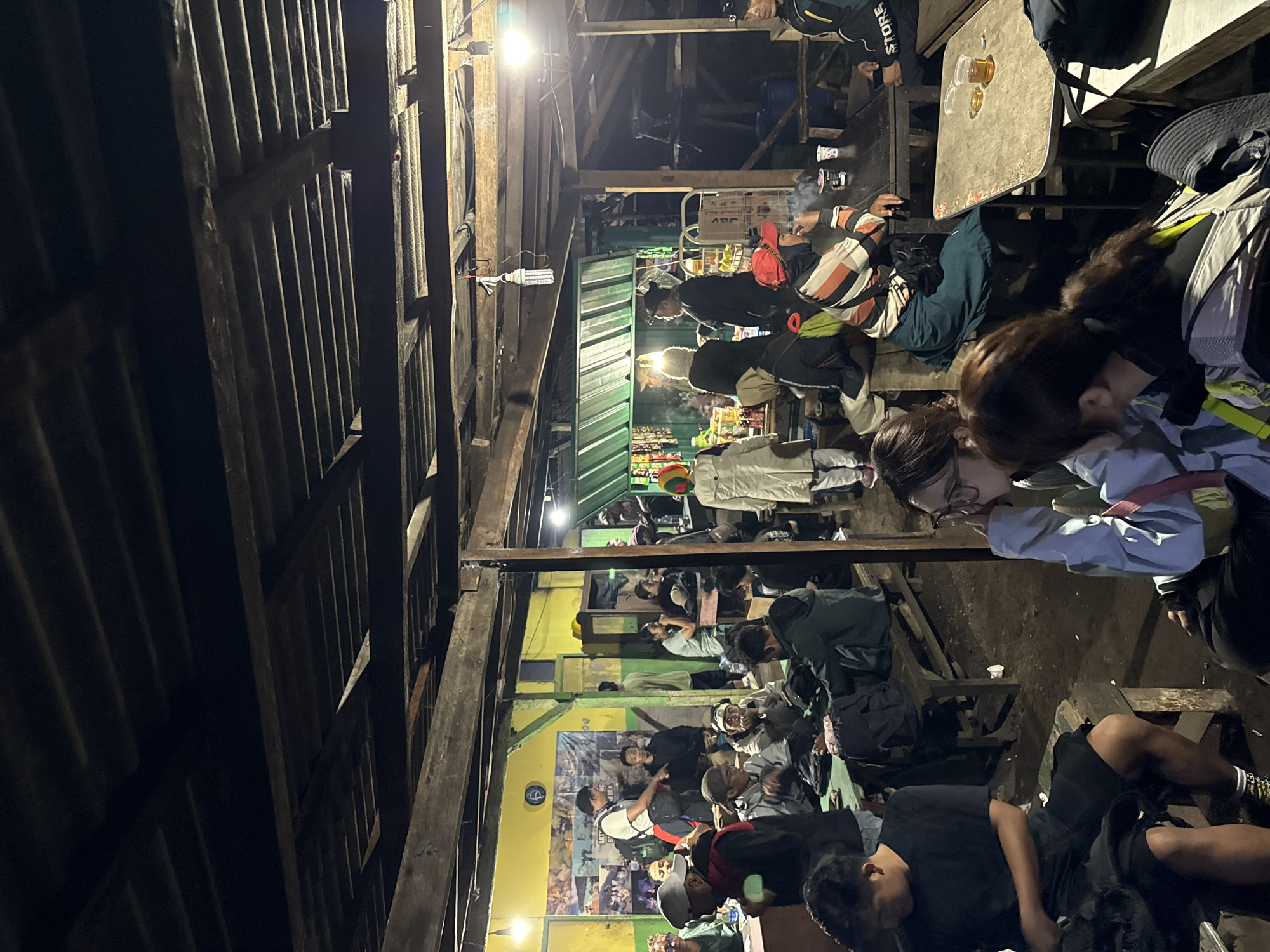 The image shows a crowded outdoor gathering in an unidentified location in Unknown, Indonesia. The space is under a wooden structure with a corrugated roof. People are seated at tables, some are standing, and at least one is lying down on a bench. Several people are smoking. The setting appears to be a restaurant or cafe. The wall in the background is painted yellow and adorned with posters. The lighting is dim.
