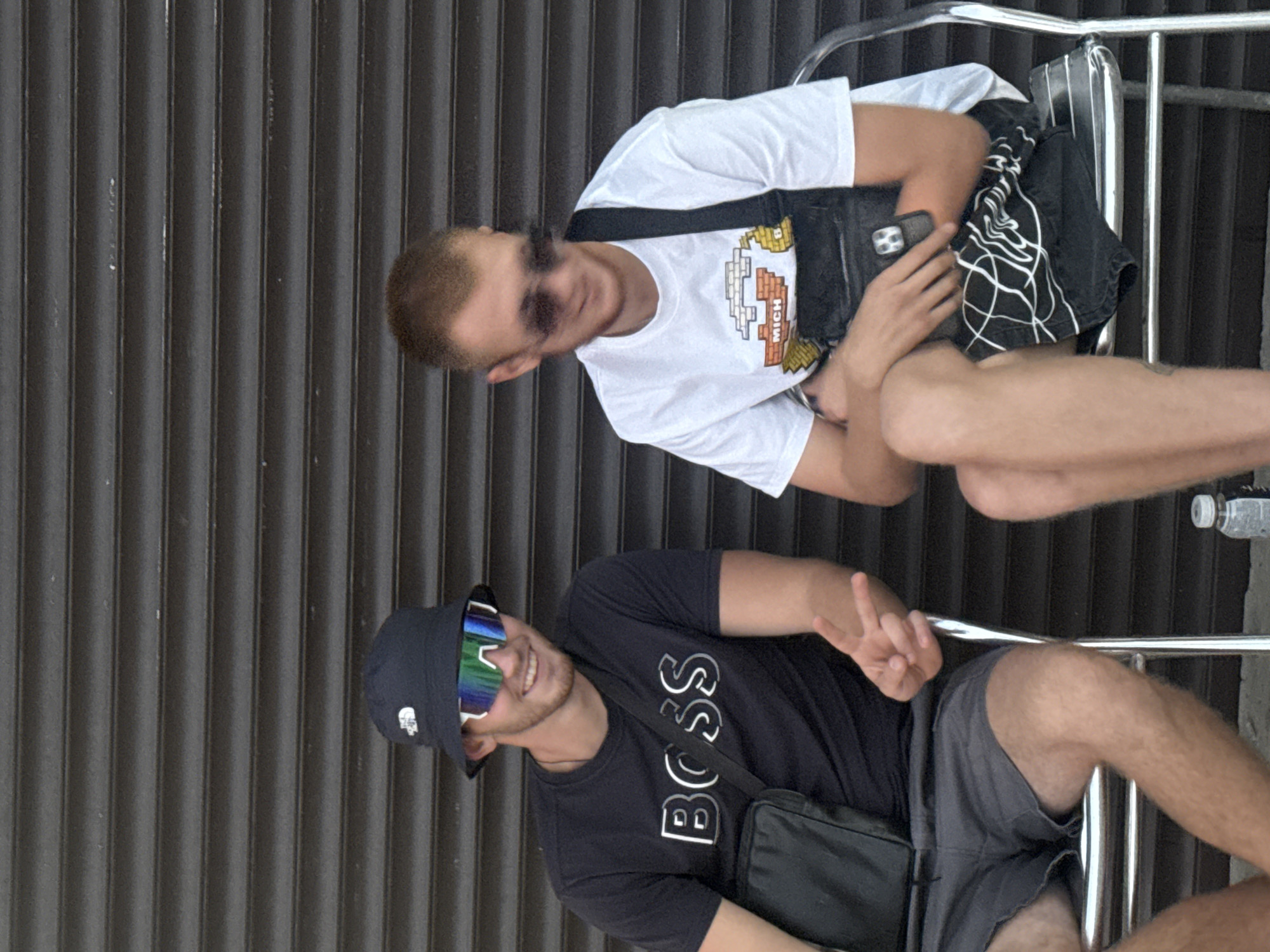 Two young men are sitting on metal chairs outdoors in front of a dark, corrugated metal wall. The man on the left wears a black bucket hat, large sunglasses with reflective lenses, and a black t-shirt with the word "BOSS" printed in white outlined in silver. He also has a black crossbody bag and is wearing grey shorts. His right hand is raised, displaying a peace sign. The man on the right wears sunglasses, a white t-shirt with a pixelated graphic design and the word "MICH" underneath, and black shorts with a white abstract pattern. He has a black fanny pack worn crossbody and is holding a black smartphone in his left hand. A plastic water bottle is visible on the ground in the lower right corner of the frame. The scene appears to be in Partenit, Ukraine.