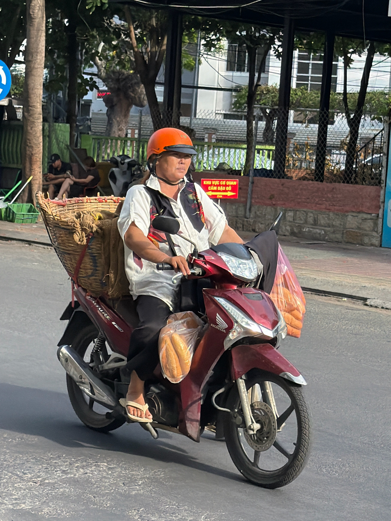 A person wearing an orange helmet and a white shirt with a colorful pattern is riding a red motorcycle in Nha Trang, Vietnam. The motorcycle has a large wicker basket strapped to the back, filled with goods, including sacks and what appears to be bread. Another bag of bread is visible on the front of the motorcycle. The person is wearing black pants and flip-flops. In the background, there are trees, buildings, and a chain-link fence. A red sign with yellow text is also visible in the background. The scene appears to be on a street in an urban area.