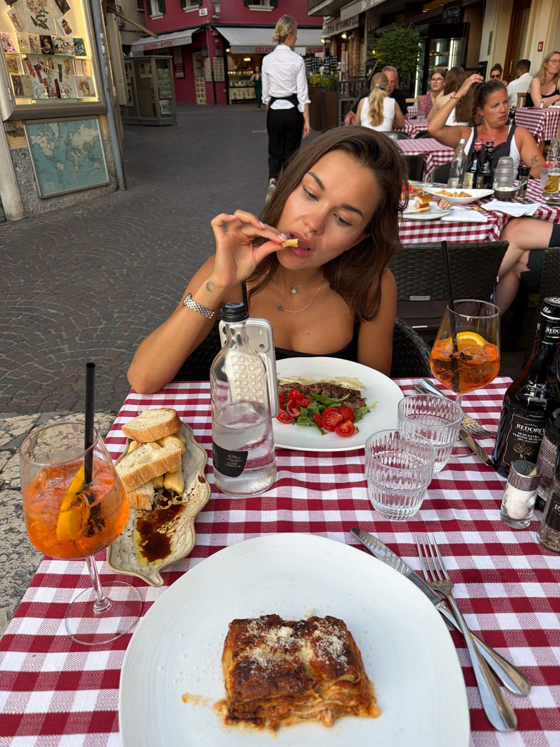 A young woman is seated at an outdoor table at a restaurant in Sirmione, Italy, eating French fries. She has long brown hair and is wearing a black tank top and a necklace. In front of her is a plate of lasagna, a glass of an orange Aperol Spritz, and a bottle of water. The table is covered with a red and white checkered tablecloth. In the background, other diners are visible, as well as shops and buildings typical of an Italian town. The scene is illuminated by daylight.