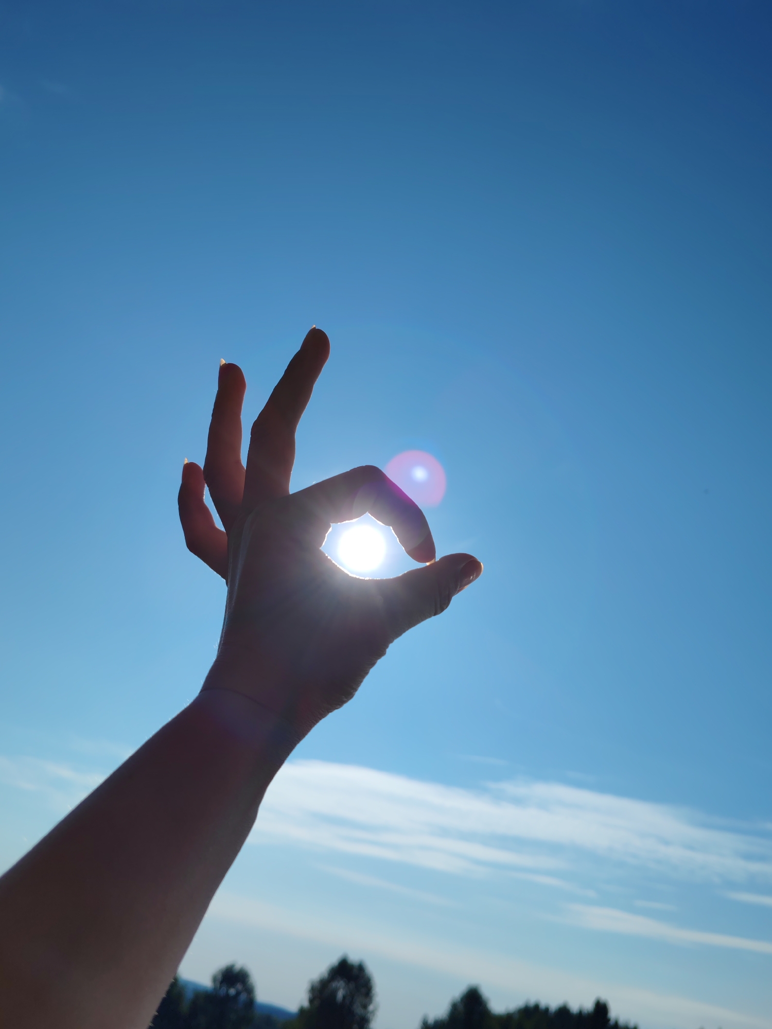 A human arm and hand are silhouetted against a bright blue sky. The hand is making an "okay" gesture, with the thumb and index finger forming a circle, and the other fingers extended upwards. The sun is visible through the circle formed by the fingers, creating a lens flare effect. In the distance, the silhouettes of trees and hills are visible against the horizon. The image appears to have been taken outdoors on a clear day, likely during daylight hours. No specific landmarks or indications of the city of Чухлэм, Russia are present in the image.