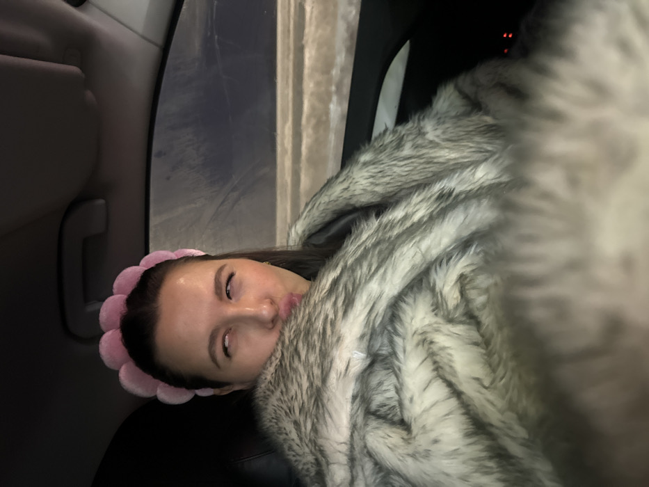 A forensic examination of the image reveals a female subject seated inside a vehicle. The specific city location of Akseno-Butyrki, Russia, is not discernible from the provided visual cues within the car's interior. The woman, with dark hair, wears a light pink, plush, scalloped headband. Her eyes are rolled upwards, exposing the sclera, and her lips are pursed in an exaggerated pucker. She is covered from the chest down with a thick, shaggy faux fur blanket or coat, displaying a mottled pattern of light gray, white, and dark tones. The upper interior of the vehicle, including a gray headliner and a grab handle, is visible. Through the passenger window, a dark, blurry exterior environment with faint horizontal streaks of light is observed, suggesting a nighttime setting or motion. The overall lighting within the vehicle is dim.