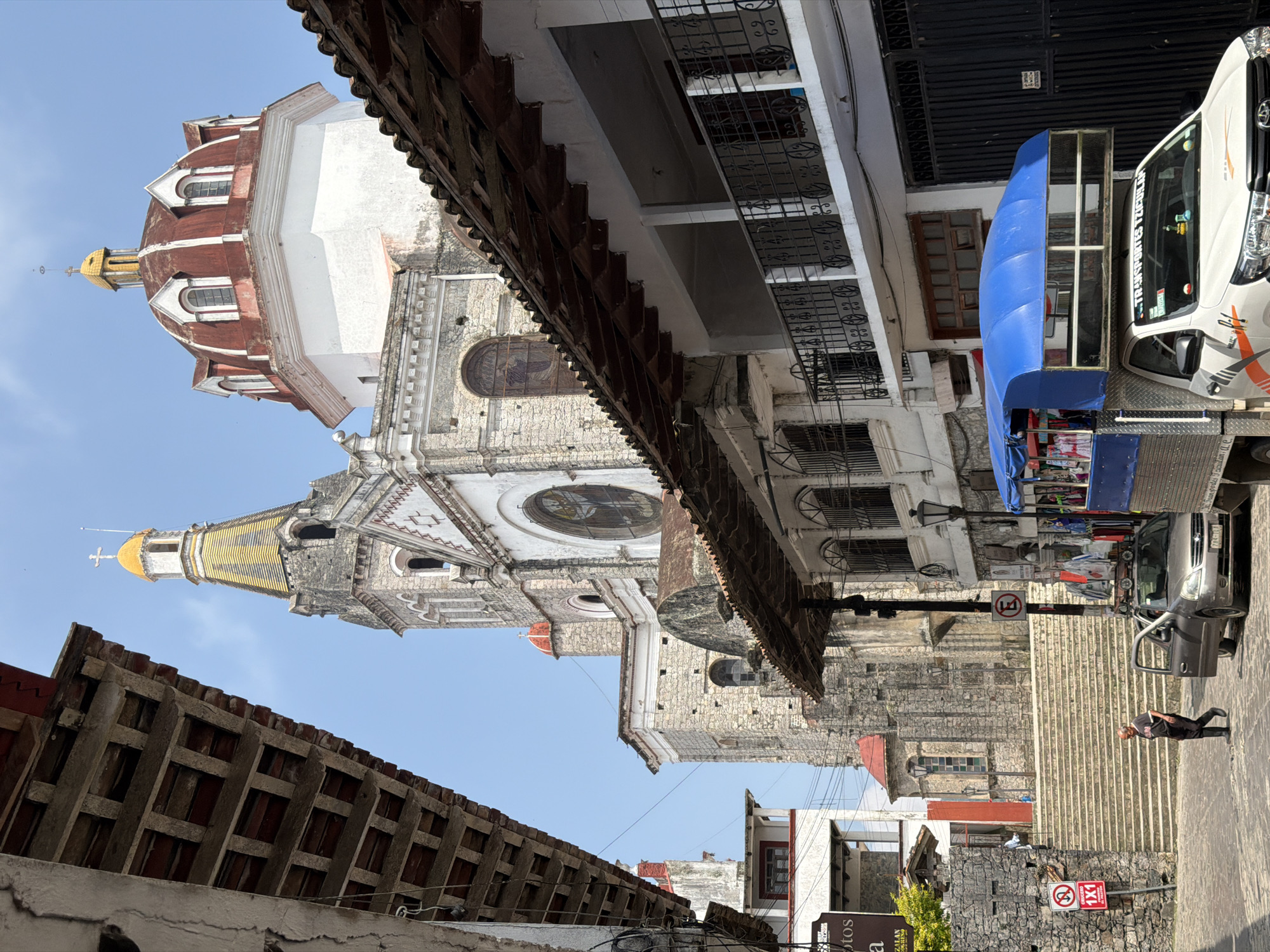 A low-angle, rotated image shows a street scene in Cuetzalan, Mexico. The prominent feature is a large, ornate Catholic church built of stone. The church has a tall, square bell tower with a yellow-tiled roof and a cross on top, and a large dome with a red roof. The facade features arched windows and intricate carvings. The street is lined with buildings, some with balconies and wrought-iron railings. A blue-covered transport vehicle is parked on the right side of the street, near a flight of stairs. A person is visible walking down the stairs. Various signs and urban objects are present along the street. The sky is clear and blue.