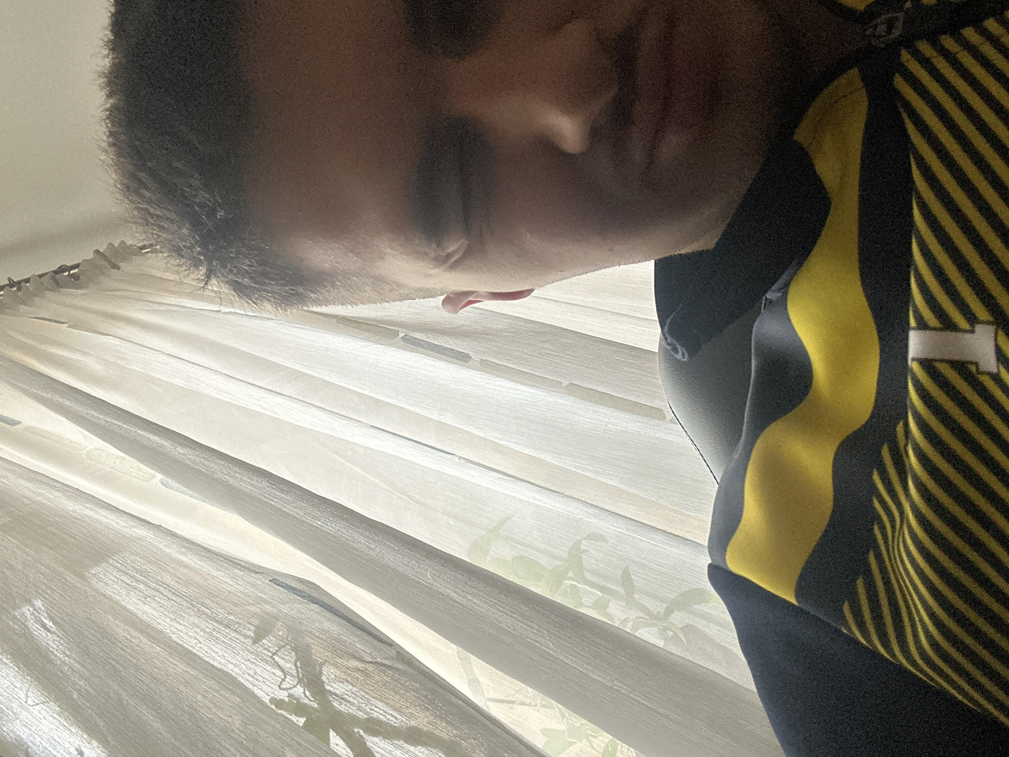 A close-up, low-angle shot of a person indoors. The person has short, dark hair and their face is partially visible with their eyes closed. They are wearing a garment with black and yellow stripes. Behind the person is a window covered with white, vertical blinds. The view through the blinds suggests a bright, outdoor environment with some foliage visible. The location is not discernible from the given image to be in Sofia, Bulgaria.