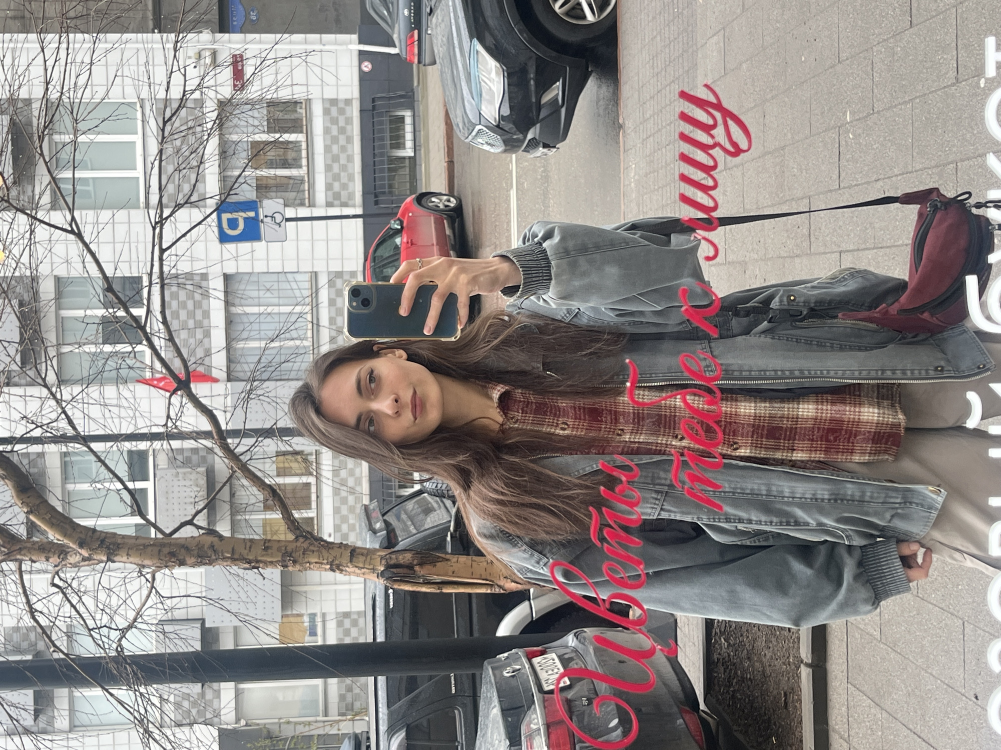 The image depicts a young woman holding a smartphone up in a mirror taking a selfie. She has long brown hair and is wearing a red and brown plaid shirt under a gray denim jacket. A red purse is slung over her right shoulder. The mirror reflects a street scene. Several cars are parked on the side of the road and buildings are visible across the street. The reflective text "Любимой теще к 8-му Марту" is written across the mirror. The location appears to be in Krasnoyarsk, Russia.