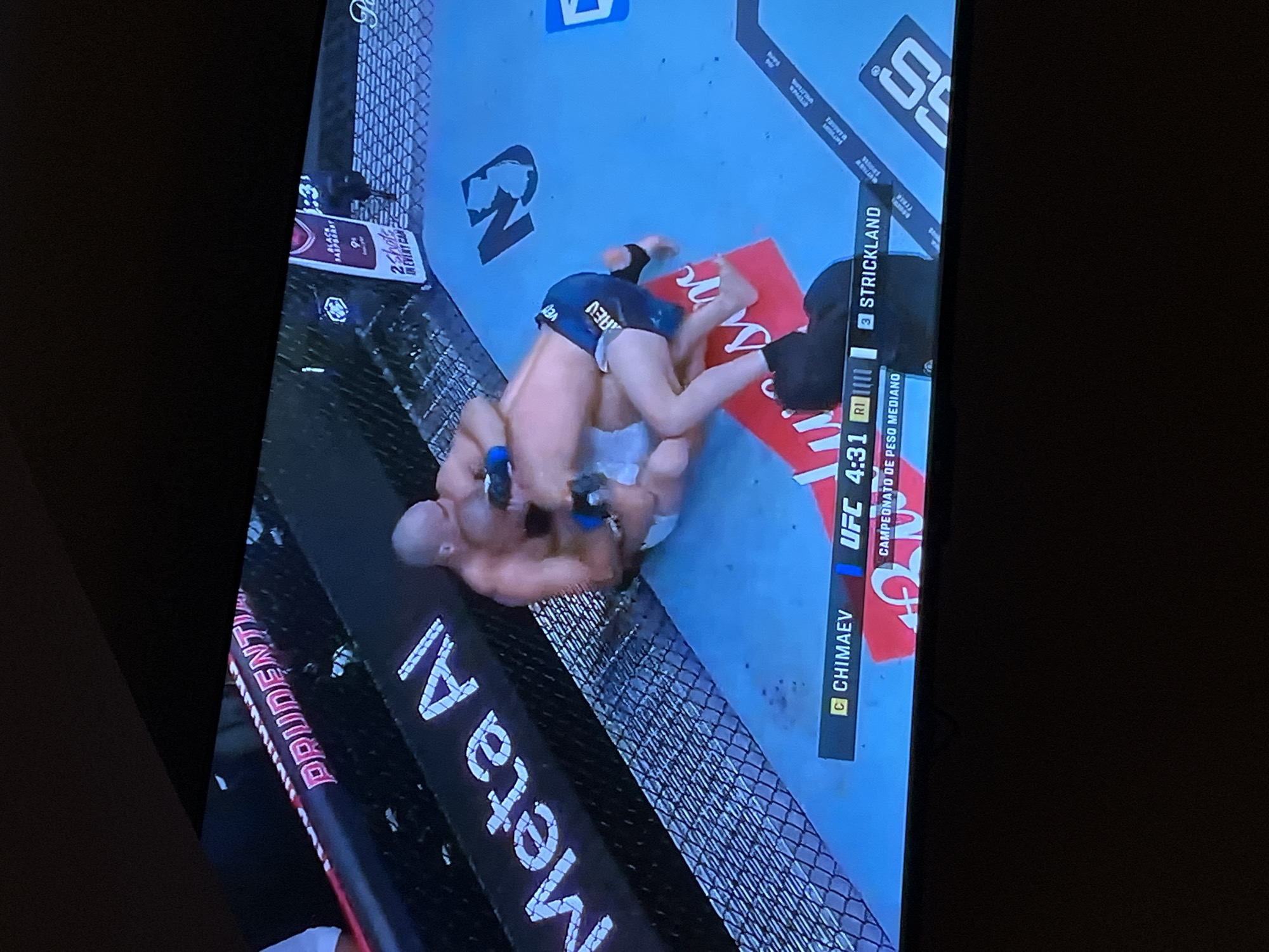 The image depicts a UFC match, with fighters Chimaev and Strickland engaged in grappling on the mat. The environment is the fighting cage, surrounded by sponsor logos such as "Meta AI" and "PRUDEN". A corner of the cage shows advertisements for "2 shots IN EVERY CAN". The UFC broadcast overlay displays the time as 4:31 of round 1, with information in Spanish indicating a middleweight championship. The scene appears to be a broadcast captured on a television screen. There is no indication of location in San Bernardino Tlaxcalancingo, Mexico.