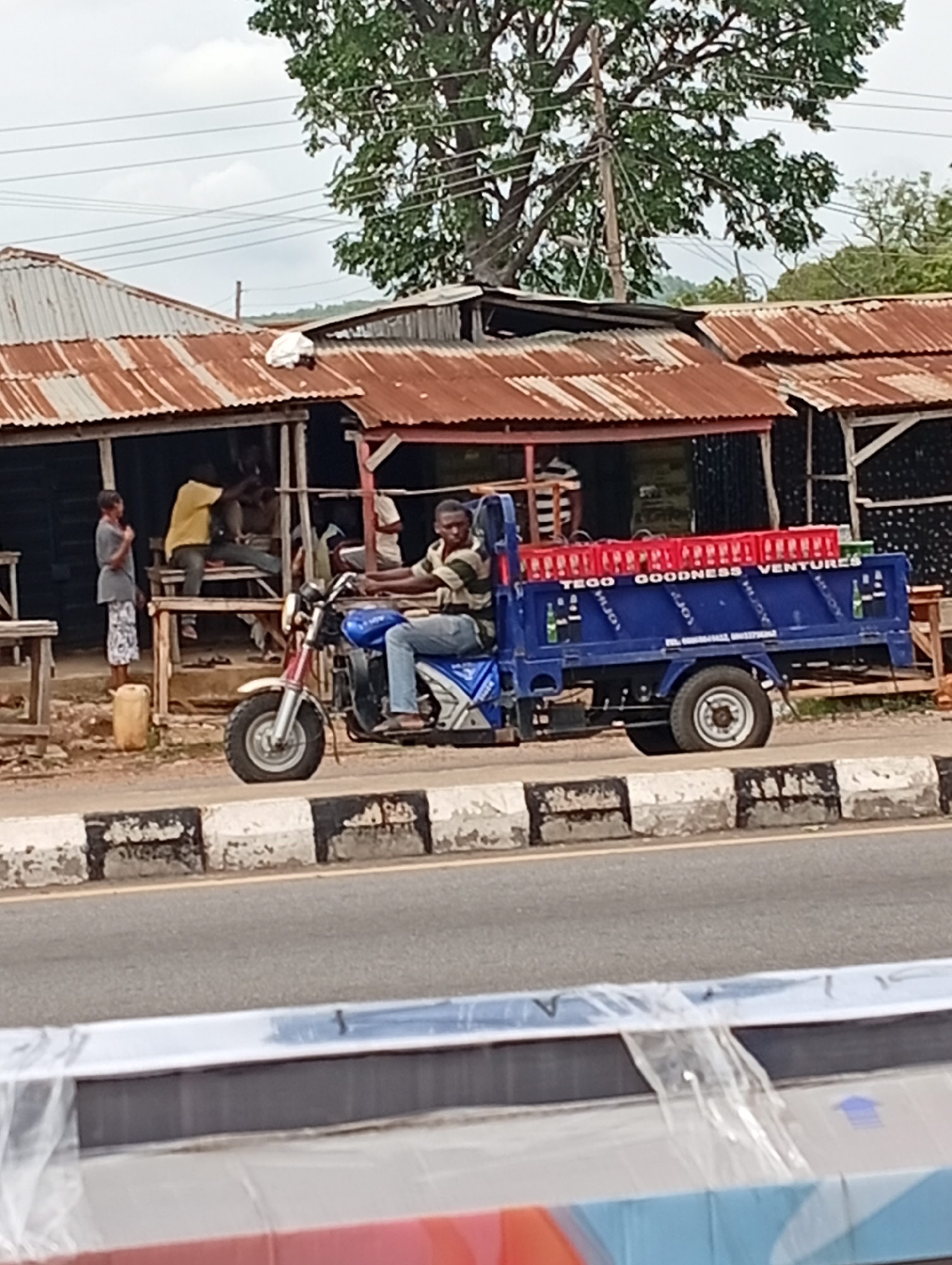 In Adankolo, Nigeria, a man rides a blue three-wheeled motorcycle truck along a road with a black and white curb. The truck bed is filled with red crates of what appear to be drinks. The side of the truck reads "TEGO GOODNESS VENTURES." Behind the truck, a group of men are gathered outside of a row of single-story buildings with rusted corrugated metal roofs. One man stands and looks toward the truck. A tree is visible in the background. In the foreground is a blurred object wrapped in plastic.