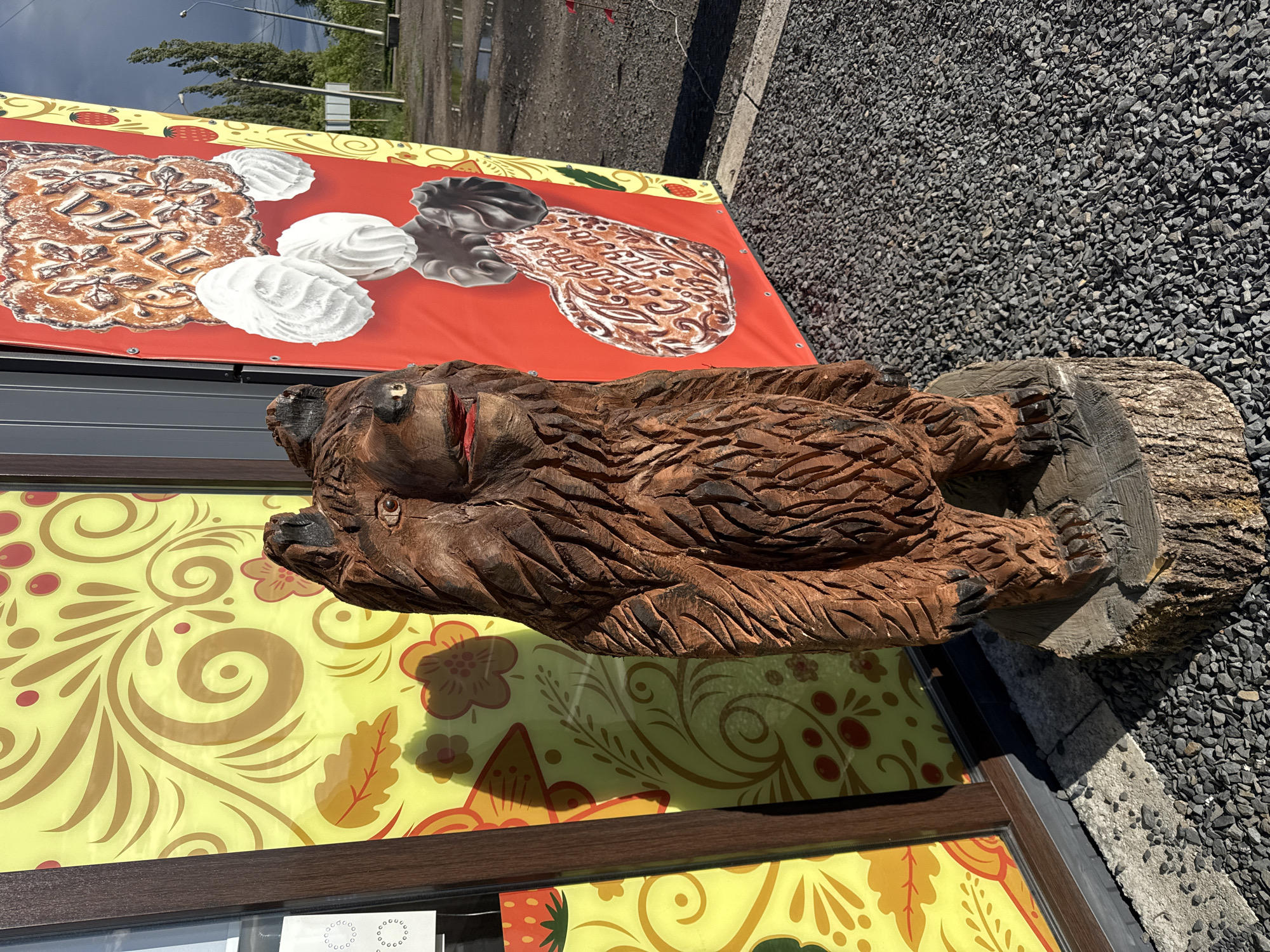 A wooden bear sculpture sits atop a tree stump beside a building decorated with dessert-themed banners. The location is Novaya Usman, Russia. The foreground consists of gravel.