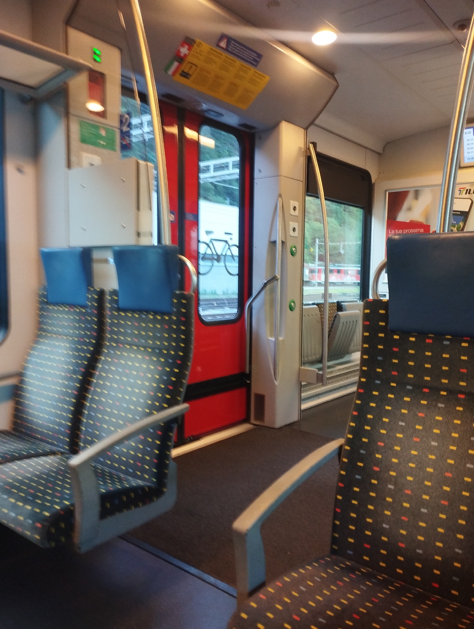 The image captures the interior of a train carriage. Two seats with patterned upholstery are visible in the foreground, one on the left and one on the right, both facing forward. The seats have blue headrests and grey armrests. To the left, a red door with a window is visible, and beyond it, a window shows a green, tree-covered hillside. A bicycle graphic is visible on the exterior of the train through the window. In the center of the carriage, a vertical silver pole and a grey handrail are present. Various signs and lights are mounted on the wall. The floor is covered with dark carpet. In the background, a digital display shows departure times, and a red and white advertisement with Italian text is posted. The overall impression is of a clean and functional train interior.