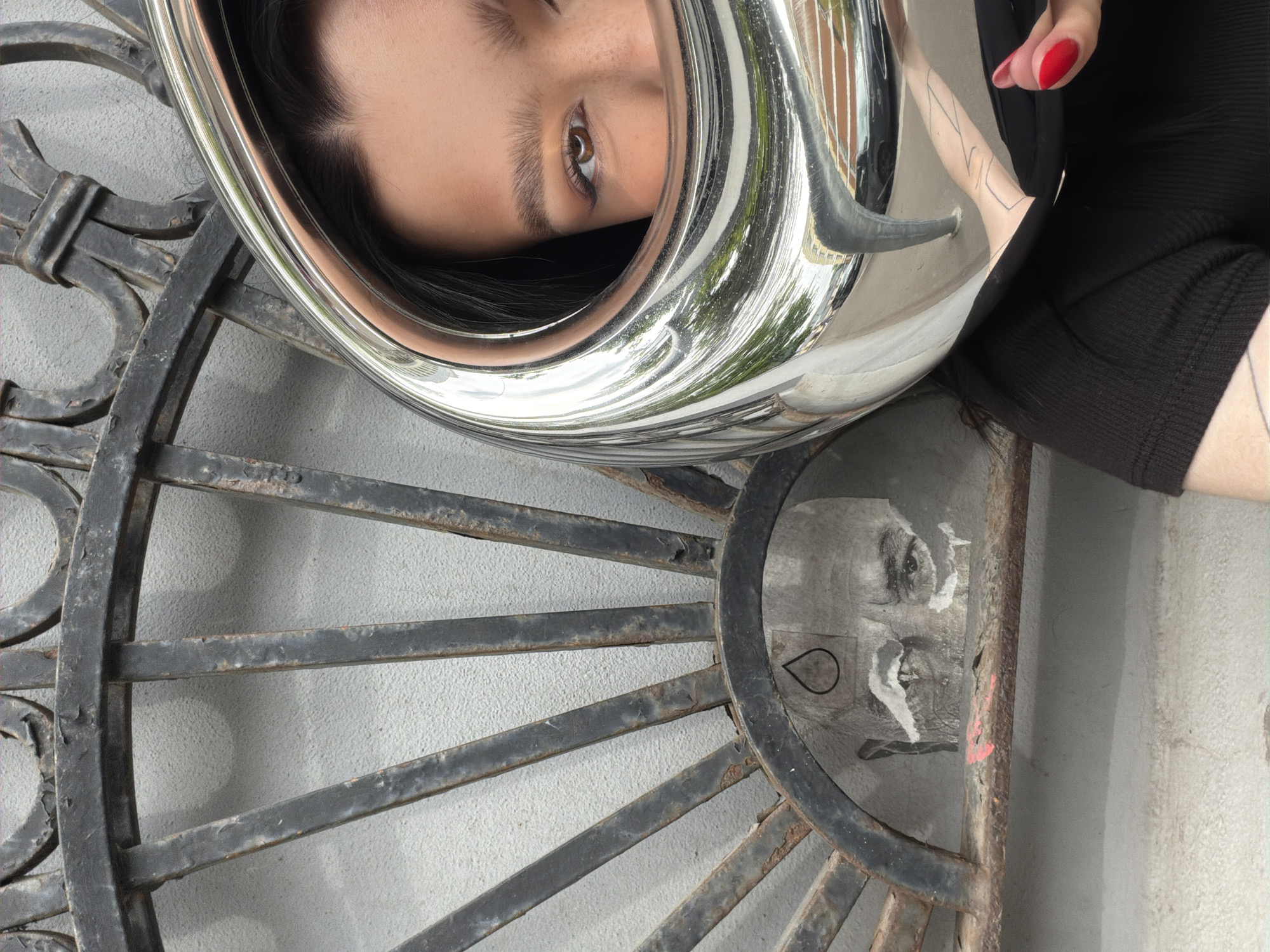 The image presents a reflective surface capturing a face with an eye looking directly at the viewer. The surface resembles a silver metallic object, possibly part of a musical instrument or a decorative piece. A hand with red nail polish is visible on the upper right side of the reflective surface. The background consists of a black metal structure, seemingly a decorative wheel or grate, set against a weathered white wall. A ripped poster or sticker with a face is visible through the metal grate.
Based on the reflection and the overall aesthetic, this image may be shot in Moscow, Russia. This is based on the style of the architecture and the design of the surrounding metal structures.