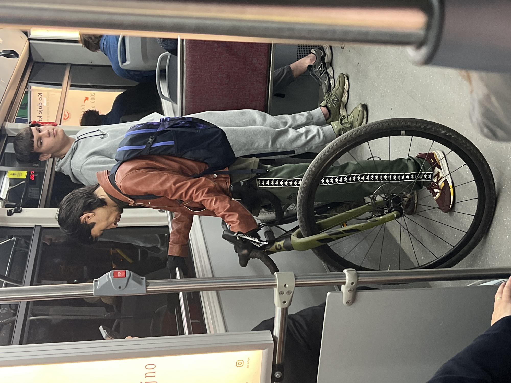 The image depicts an interior scene on what appears to be a bus or tram. A person wearing a brown jacket and green pants is standing next to a bicycle. Another person in a grey hooded sweatshirt and grey sweatpants is lying down on the floor. There are other people present in the background, one seated. The overall impression is one of a crowded public transportation setting. The image does not provide explicit confirmation that the location is within the city of Palilula Urban Municipality, Serbia, but the presence of Cyrillic text on the bus signs implies it is somewhere in Serbia.