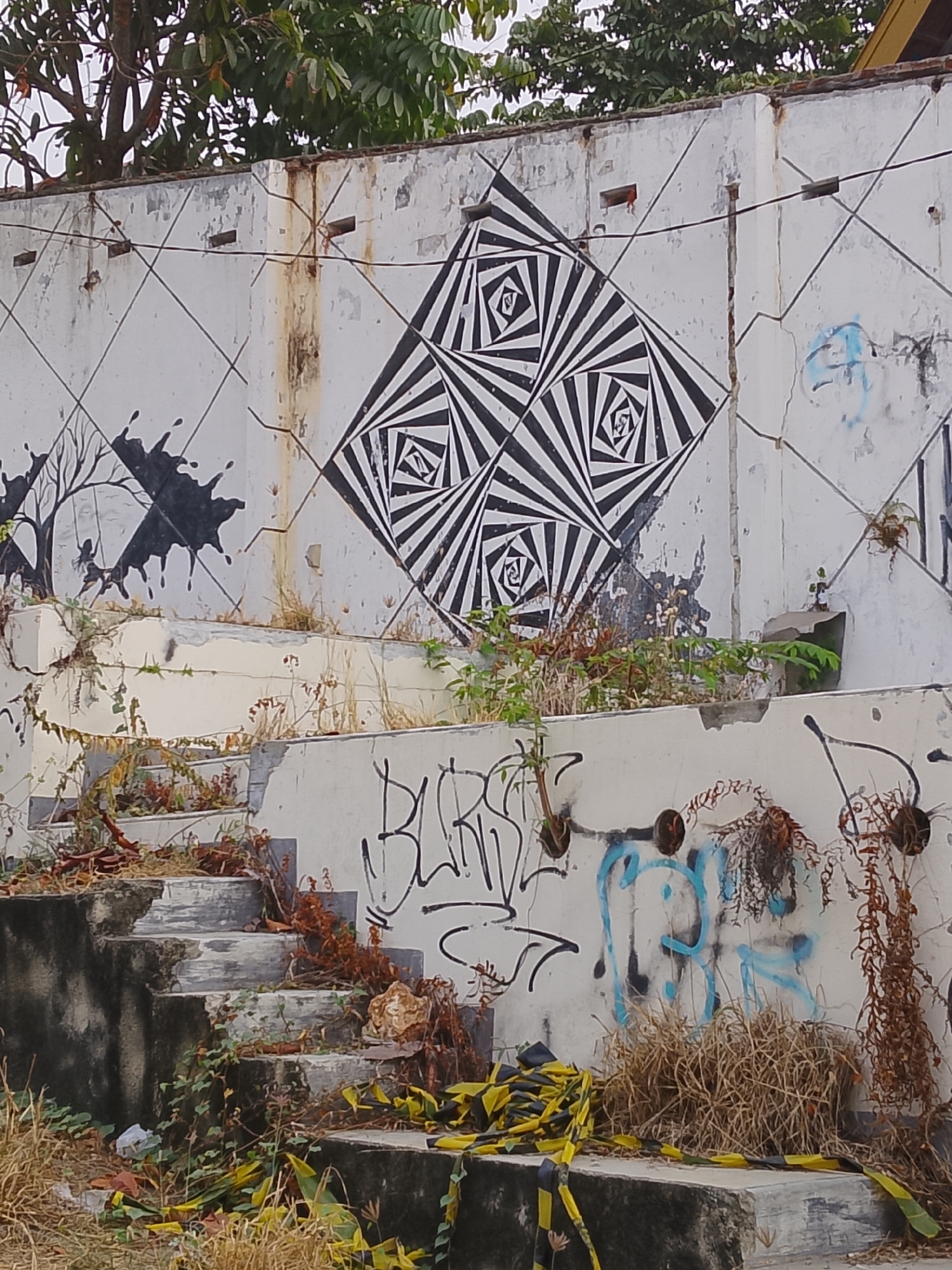 In Ngelo, Indonesia, a dilapidated concrete structure stands covered in graffiti and overgrown with vegetation. The wall, once painted white, is now peeling and stained, with evidence of rust streaks running down from the top edge. A large, optical illusion-style mural of black and white geometric shapes dominates the upper portion of the wall. To the left, a silhouette of a tree with splattered ink marks is visible. Below the wall, a set of worn concrete stairs, also encroached upon by weeds and dried leaves, descends from left to right. The ground area around the stairs is littered with debris, including crumpled white paper and yellow and black caution tape. Further to the right, more graffiti adorns the wall, including blue scribbles and black abstract lines. Dry, brown grass and hanging vines add to the sense of neglect and decay. The scene is lit by an overcast sky, casting a soft, diffused light.
