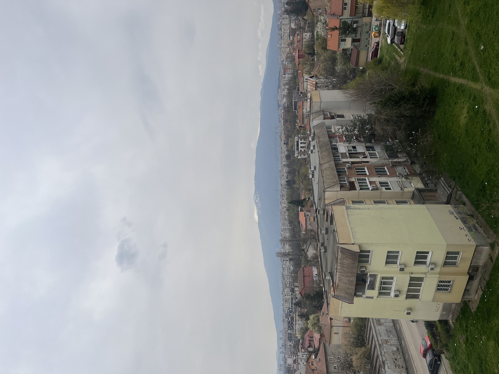 The image presents a landscape view of Sofia, Bulgaria, under a cloudy sky. A mountain range is visible in the background. The foreground features residential buildings, including a pale yellow structure with multiple windows and air conditioning units. Nearby is a street with parked cars. Green grass is on the right.