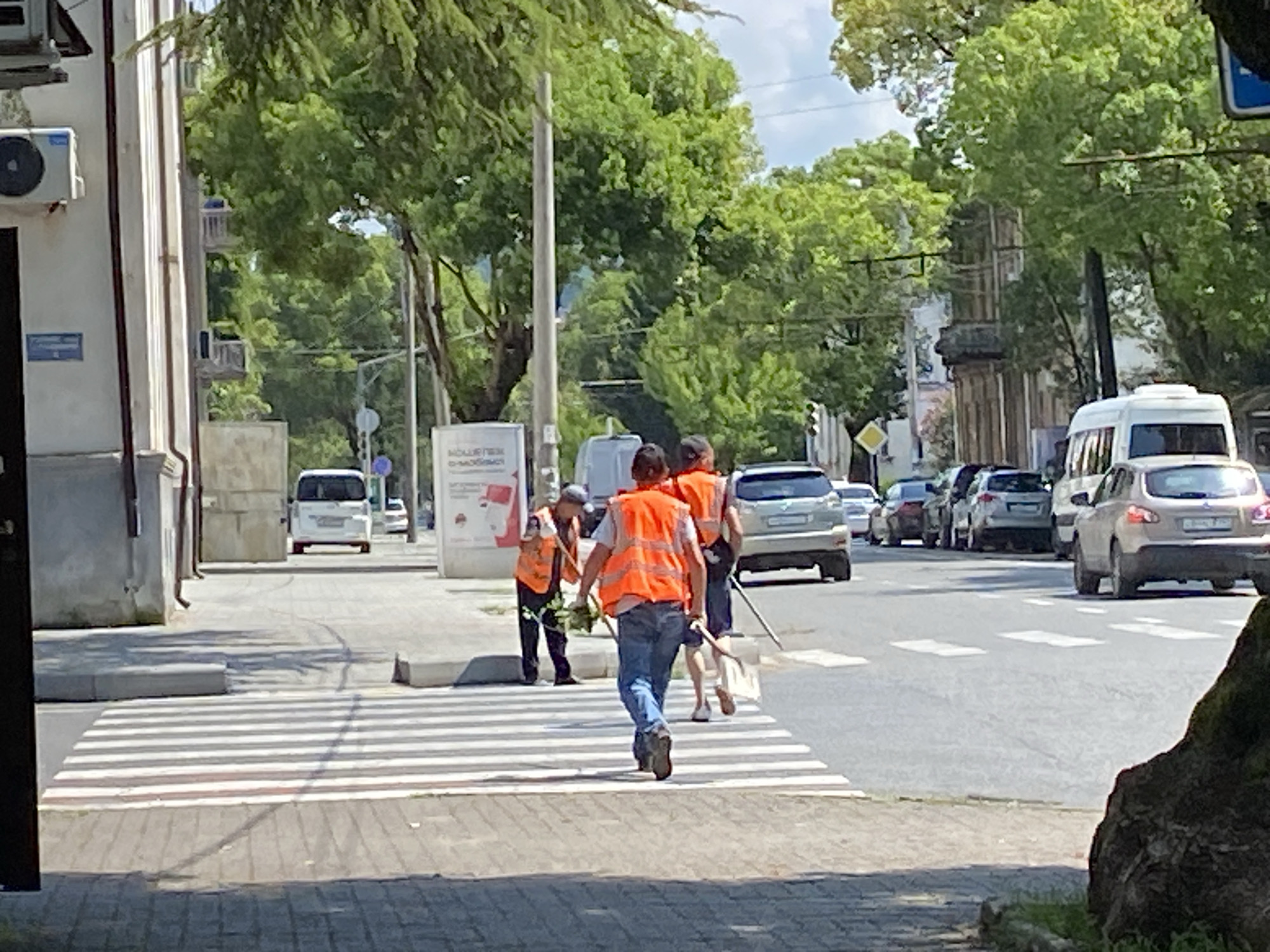 A group of street cleaners, wearing orange safety vests and blue jeans, are crossing a pedestrian crossing on a street in Sokhumi, Abkhazia. The cleaner in the foreground is walking away from the camera, carrying a broom. To their left, two other cleaners are walking side-by-side, one holding a broom and a plant. Cars are visible on the street, and buildings and trees line the sidewalk. The scene appears to be on a sunny day.