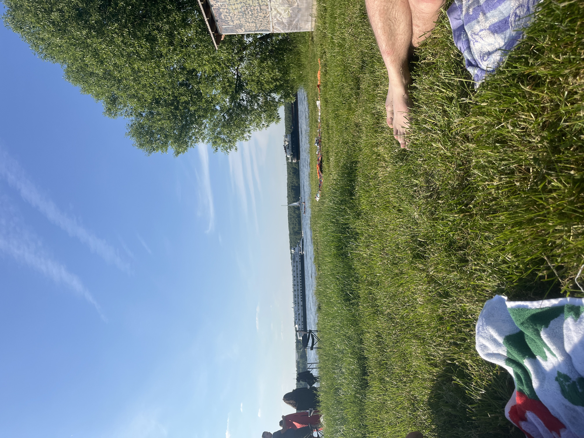 The image presents a daytime scene on a grassy riverbank in Kapustino, Russia, observed from a low perspective. The sky is bright blue with minimal, thin white cloud formations. A wide river occupies the midground, featuring a large white multi-deck passenger ship on the left, a smaller white sailboat in the center, and a dark-hulled cargo vessel further right, all moving along the waterway. A dense treeline marks the distant opposite bank of the river.
In the midground on the left, two to three individuals are seated in portable chairs, facing the river. One person wears a black top, and another is partially obscured by a red garment. On the right side of the midground, a large tree with dense green foliage stands prominently. Adjacent to the tree is a small, rectangular shed-like structure, its exterior covered in a green and brown camouflage-patterned material.
The foreground is dominated by bright green grass. On the immediate right, the bare feet and lower legs of a person are visible, resting on the grass, with a blue and white striped towel partially beneath them. On the immediate left, a white towel with red and green patterns is laid out on the grass. Sunlight casts clear shadows, indicating a bright, clear day.