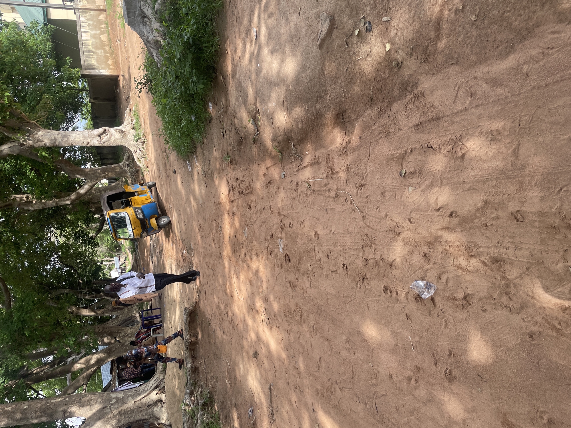 In Jalingo, Nigeria, a dusty, unpaved road passes between large trees. A yellow and blue auto-rickshaw is parked on the right side of the road. A man in a white shirt and black pants walks away from the camera on the left side of the road. To the left of the road, a person in a colorful patterned outfit walks towards the camera, and another person sits at a table. The ground is covered in dirt with tire tracks and scattered debris, including a plastic bottle. Lush green foliage grows on the right side of the road, and a concrete wall and building are visible in the background.