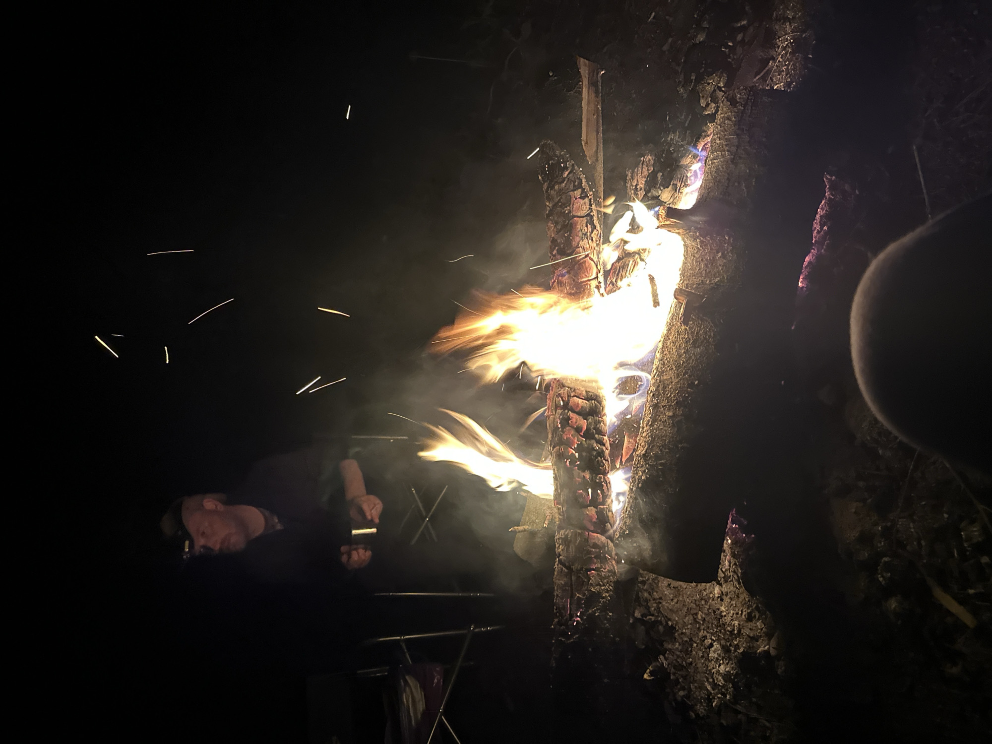 The image depicts a campfire at night. The fire is contained within a structure made of logs, with flames and smoke rising upwards. To the left of the fire, a person wearing a headlamp is holding an object. A folding table is partially visible in front of the person. Sparks are flying off from the fire, and the surrounding environment appears dark. The location is Юзуфоўскі сельскі Савет, Belarus.