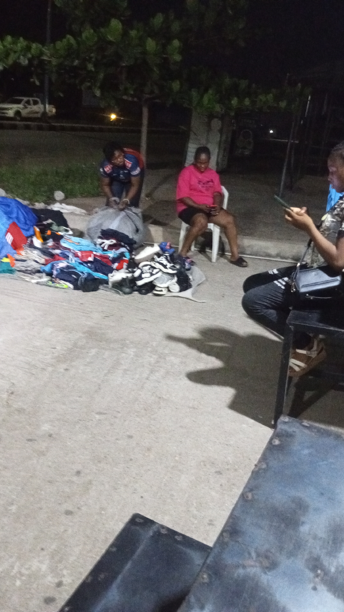 Three women are outdoors at night in Adankolo, Nigeria. One woman is seated in a white plastic chair, looking at a mobile phone. Another woman is kneeling and handling a large grey bag containing clothing. The third woman is seated in a chair, looking at a mobile phone. A pile of clothes and shoes is laid out on a white sheet on the ground. A car is visible in the background, and a large tree. A dark table or shelf is visible in the foreground. The ground is concrete.