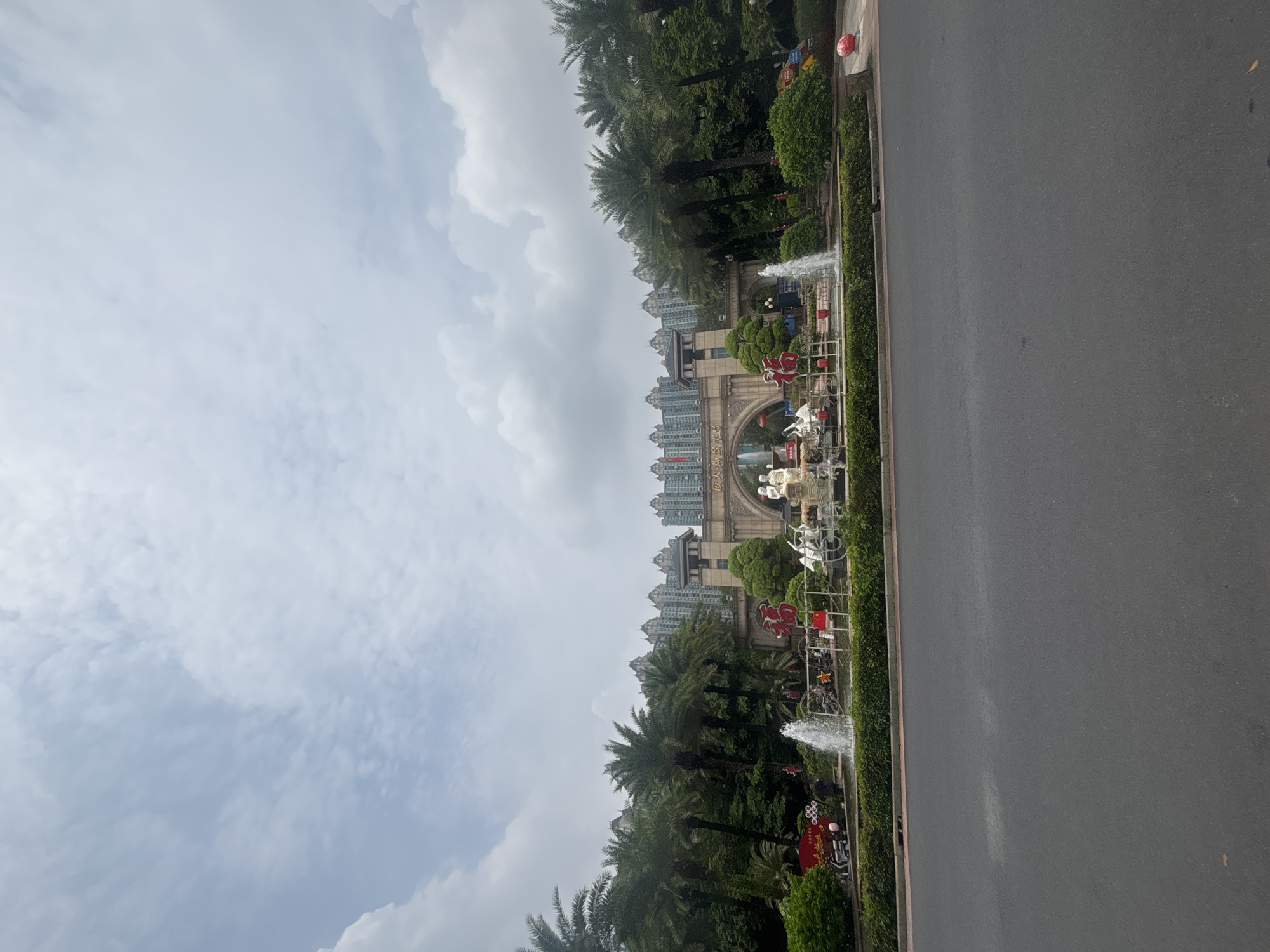 The image captures a street view in Nanhai District, China, featuring an elaborate entrance with a prominent archway. Flanking the archway are tall palm trees and lush green foliage, with modern apartment buildings visible in the background. In front of the archway, a decorative fountain is active, with water jets spraying upwards. The area is adorned with various decorations, including red lanterns, white sculptures of figures and animals, and a large red Chinese character. Some people are present, appearing to be near the decorations and possibly interacting with the environment. The scene is set under a partly cloudy sky.
