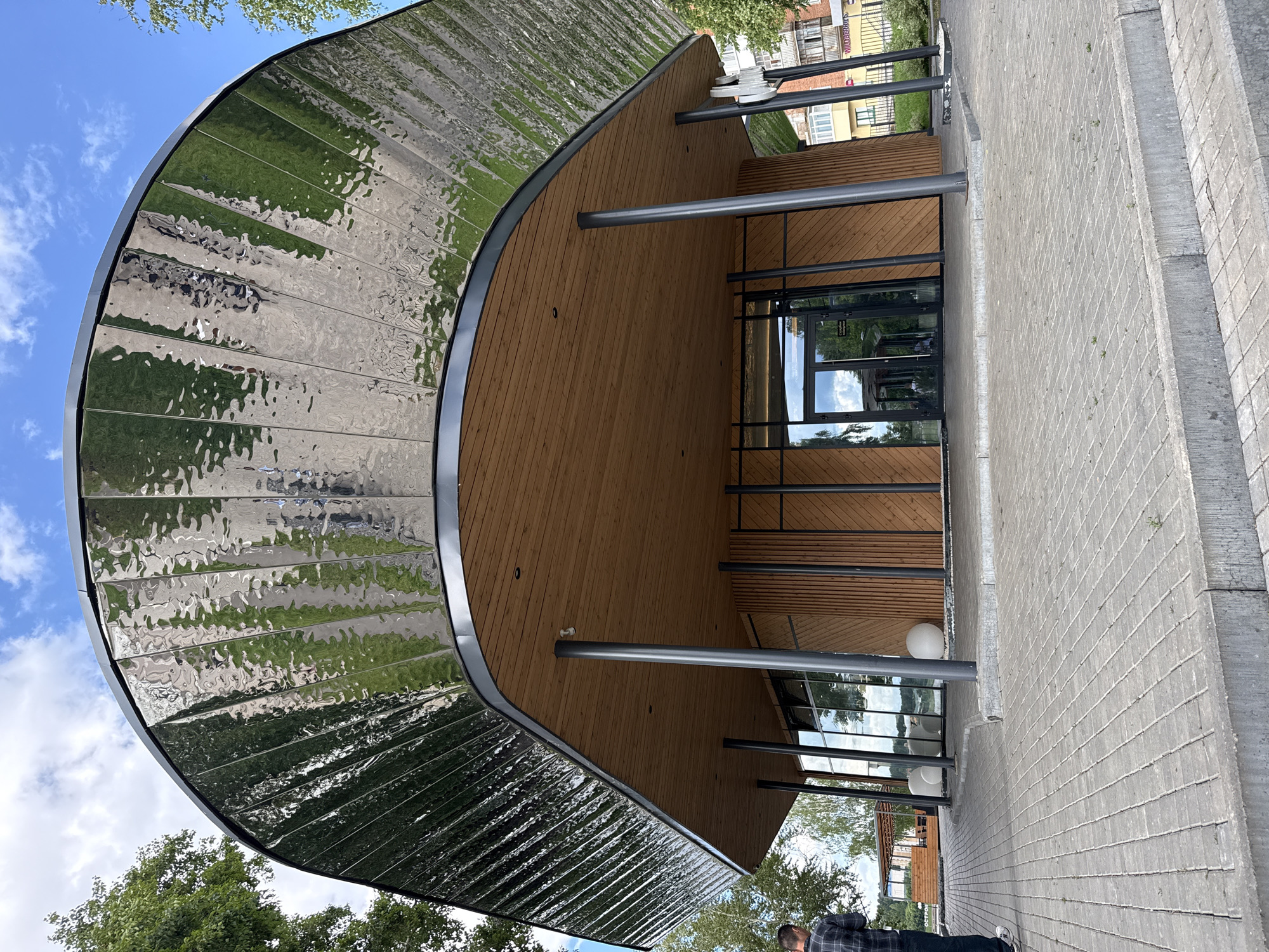 This image depicts the exterior of a modern building with a distinctive curved, metallic roof that reflects the surroundings. The building features a combination of natural wood paneling on its interior walls and a glass-fronted entrance with dark framing. Several dark metal poles support the roof, creating an open-air walkway leading to the entrance. The ground in front of the building is paved with light gray interlocking stones, and a concrete step runs along the side.
To the left of the building, lush green trees and a bright blue sky with scattered white clouds are visible, indicating fair weather and likely daytime. The metallic roof reflects the sky and greenery, creating a dynamic visual effect. In the background, a multi-story building with windows and balconies can be seen, suggesting an urban or suburban setting.
A person is partially visible in the lower right corner of the image, wearing a plaid shirt and dark pants. They appear to be walking away from the camera, towards the building. Their presence suggests the building is accessible and likely in use.
No discernible text is visible in the image, aside from what might be on signage within the reflection of the glass doors, which is too indistinct to read. The overall impression is one of contemporary architecture set within a pleasant, natural environment. The scene suggests an ordinary day, possibly late morning or early afternoon, given the brightness of the light and the clear sky. The location context is Votkinsk, Russia.