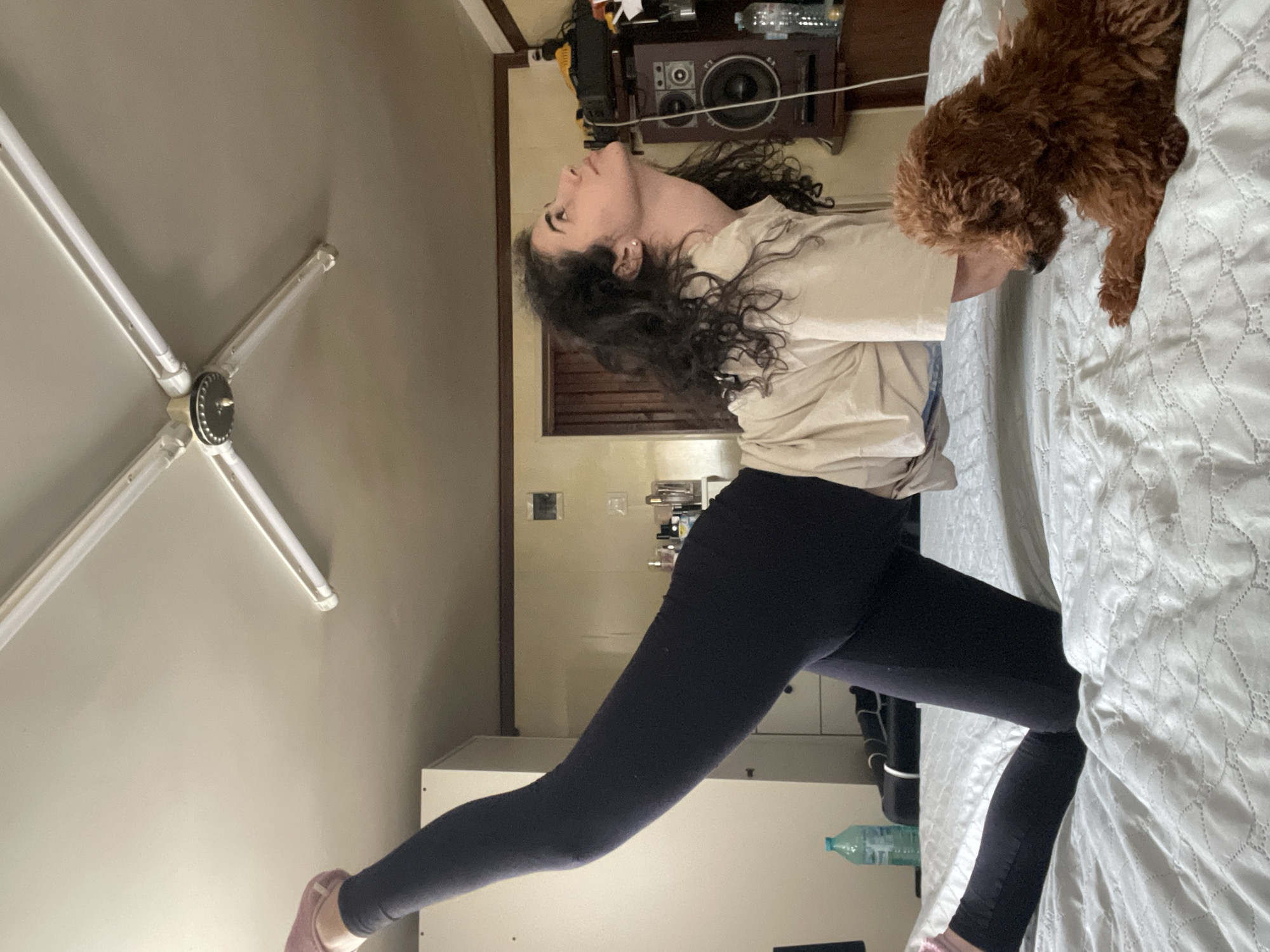 A woman is in a low lunge pose on a bed with a fluffy brown dog on it. The woman is wearing a light-colored top and dark leggings. A ceiling fan is visible in the upper left corner. A speaker and other items are on a shelf in the background. A white cabinet is near the leg that is extended backward. A water bottle is partially visible near the white cabinet. The location appears to be a bedroom; there is no evidence to suggest Sofia, Bulgaria.