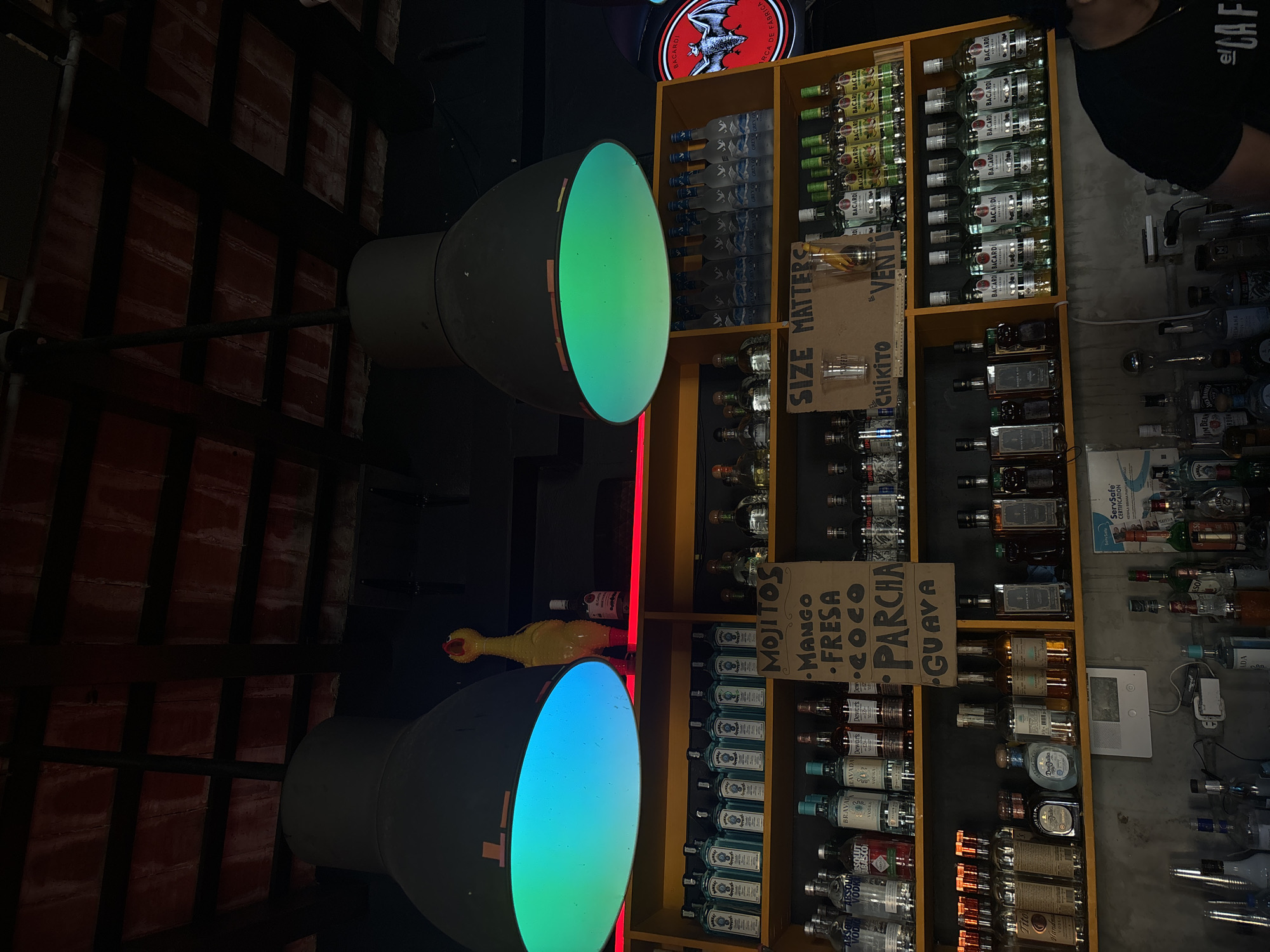 The image shows a dimly lit bar in San Juan, Puerto Rico. The ceiling is made of dark wood beams with red tiles. Two large industrial lamps hang from the ceiling, one emitting a blue light and the other a green light. A yellow rubber chicken toy is visible on a shelf behind the blue lamp.
The shelves are stocked with numerous bottles of alcohol, including vodka, gin, rum, and tequila. There are also several signs on the shelves. One sign lists "MOJITOS," "MANGO," "FRESA," "COCO," "PARCHA," and "GUAVA." Another sign reads "SIZE MATTERS."
In the foreground, a person is partially visible on the right side of the image, wearing a black t-shirt. The bar counter is made of gray concrete. Various bottles and bar equipment are visible on the counter.