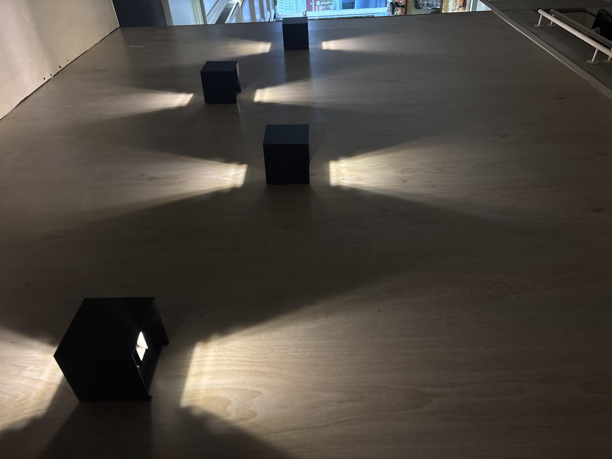 The image depicts an interior wall within a commercial establishment in Sofia, Bulgaria. The wall is constructed of light-toned wood paneling, possibly plywood or veneer, displaying prominent vertical grain. Six black, cuboid-shaped wall sconces are mounted on this surface. The sconces are arranged with two on the left side, positioned one above the other, and four on the right side, forming a diagonal ascending pattern. Each sconce projects distinct, warm-toned, conical beams of light both upwards and downwards onto the wooden wall, creating symmetrical illumination patterns. The ceiling, visible at the upper right, is an off-white or light-grey, unpainted surface, likely drywall, secured with visible dark fasteners along its edge. In the lower right foreground, a white metal handrail with multiple vertical supports is partially visible, suggesting the presence of a staircase or ramp leading downwards. Beyond the handrail, a blurred background reveals an interior space, likely a retail area, containing shelving units with items packaged in red and white, and a discernible blue light source. No individuals or specific actions are observed.