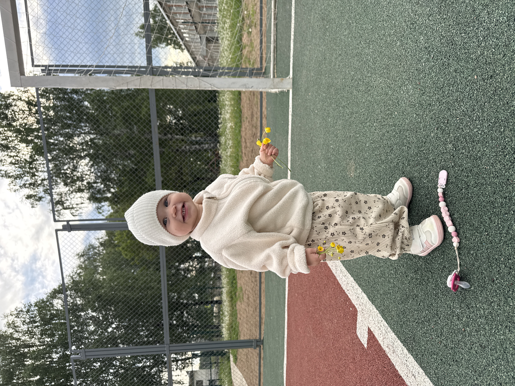 A young child is standing on a green and red surface, likely a sports court, holding yellow flowers. The child is wearing a white hat, a cream-colored jacket, and floral-patterned pants, with white and pink shoes. A pacifier is lying on the green surface near the child's feet. Behind the child is a chain-link fence, trees, and a building, with a blurry landscape visible beyond the fence. Based on the location provided (Вассята, Russia), this is a scene of everyday life in a public or residential area.