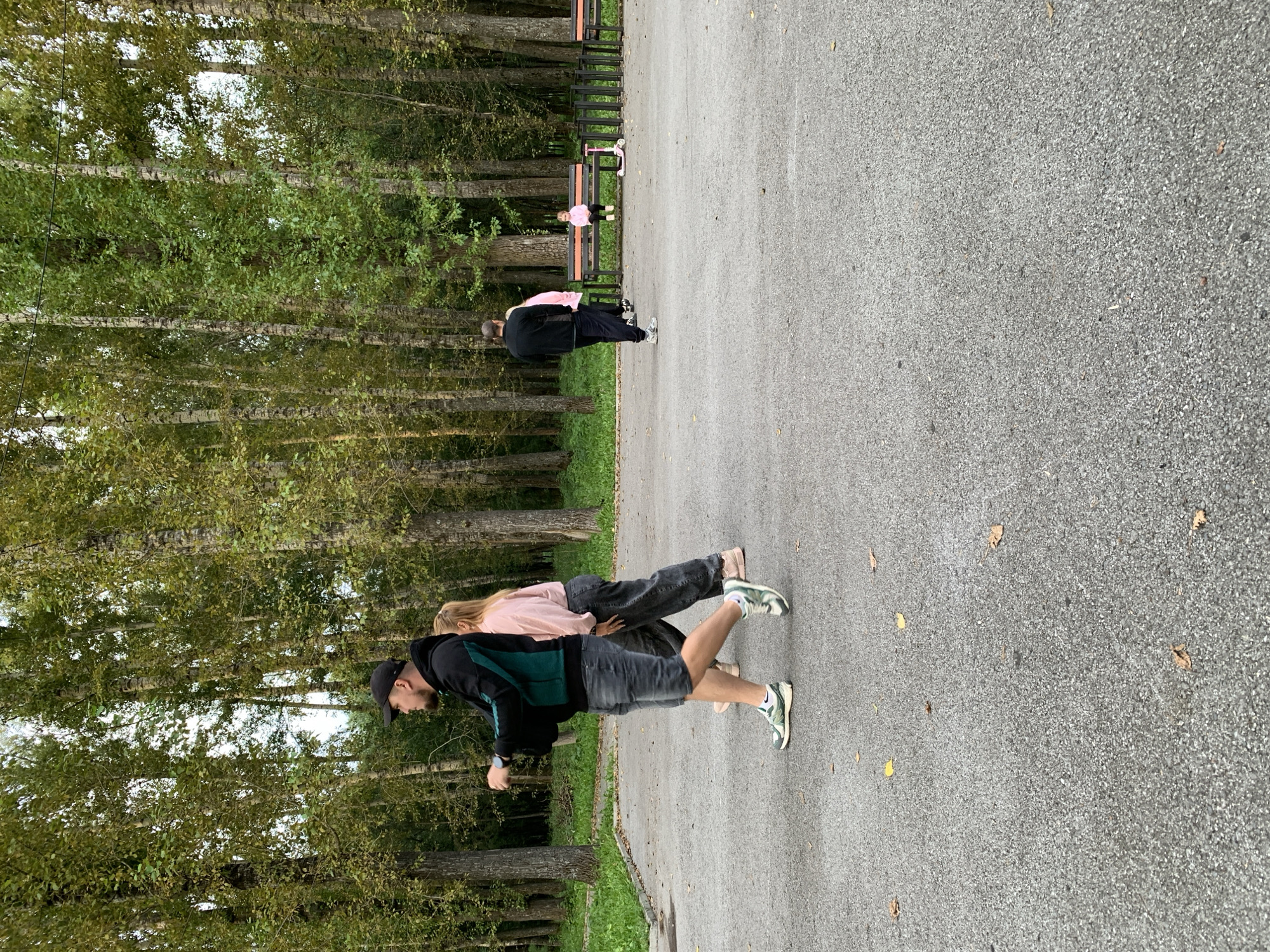 In Prokopyevsk, Russia, a paved pathway runs through a park with tall trees, some with sparse, green foliage, and others with bare trunks. A man wearing a black cap, black and green jacket, and denim shorts walks with a woman in a pink top and dark pants. Further down the path, another man in a black jacket and dark pants walks towards a bench where a young girl in a pink jacket is seated. A pink kick scooter rests on the ground beside the bench. The ground is covered with fallen leaves.